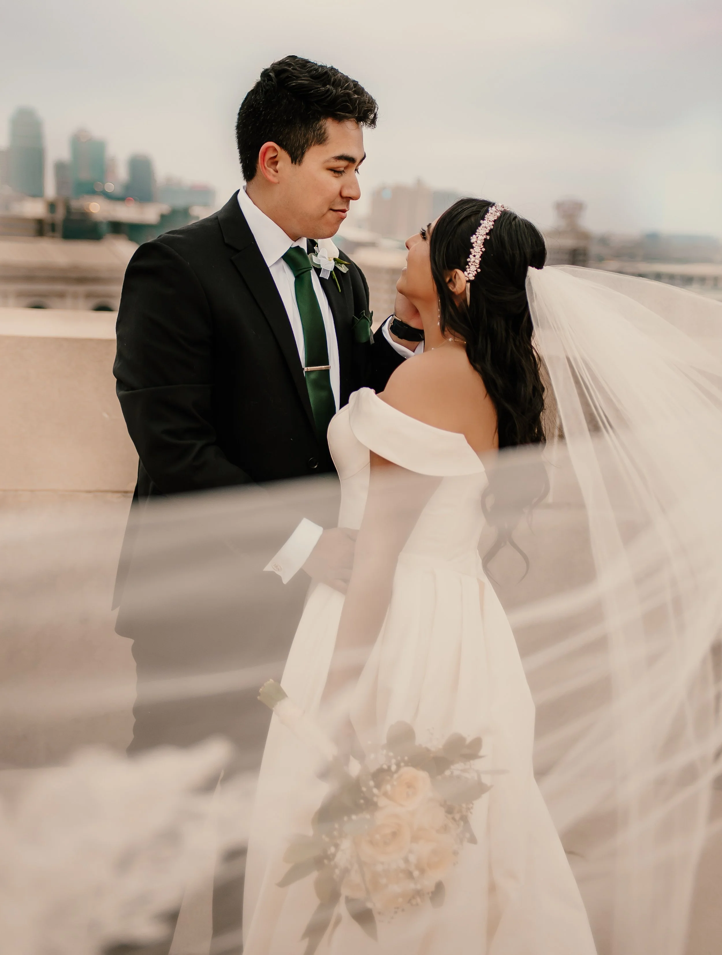 A groom and bride looking into each other's eyes on a building rooftop with city skyline in the background, the groom dressed in a black suit and green tie, the bride in a white wedding gown with a veil and headband.