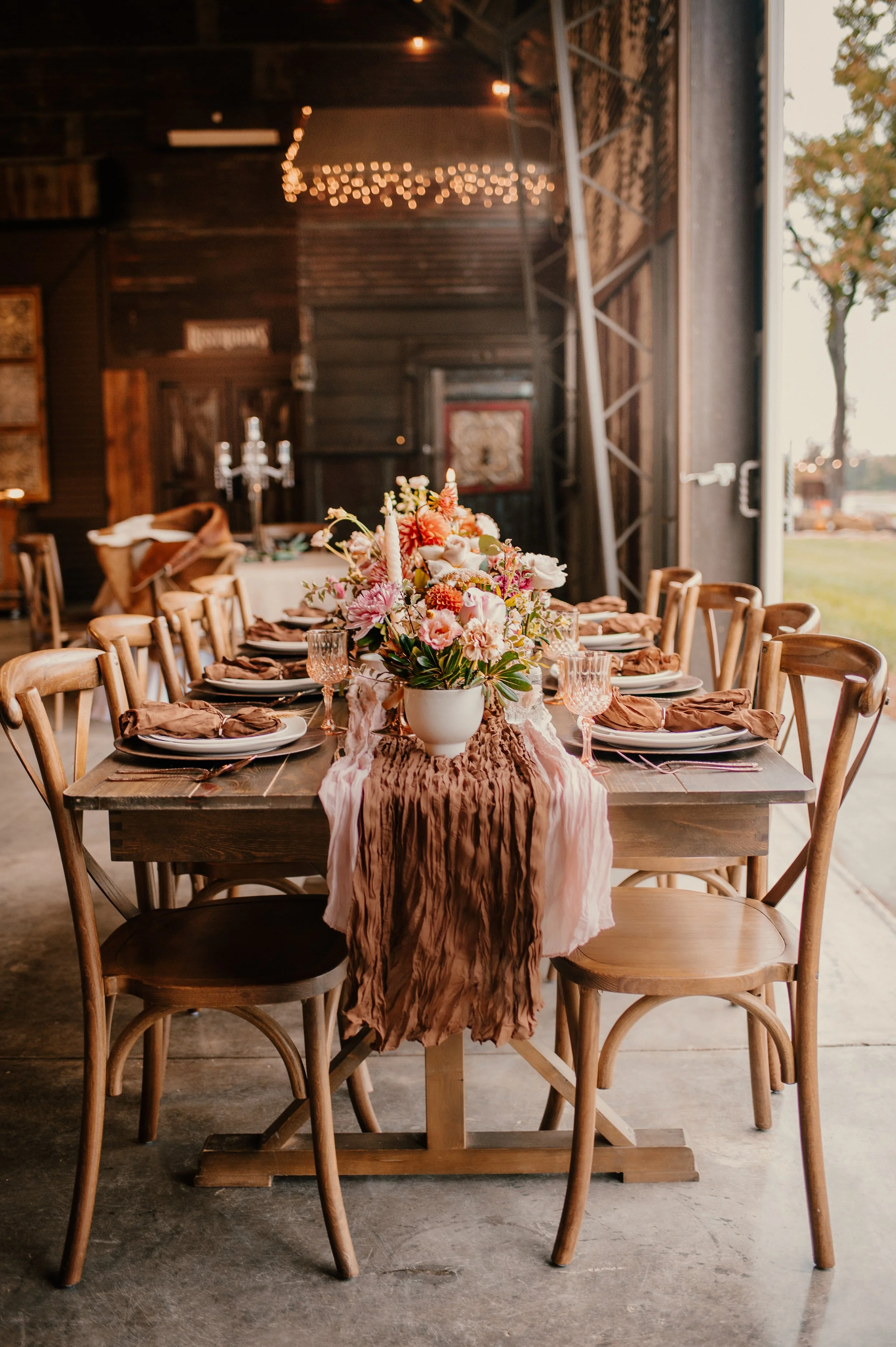 A long wooden table set for a meal with a floral centerpiece, surrounded by wooden chairs, inside a rustic barn with string lights.