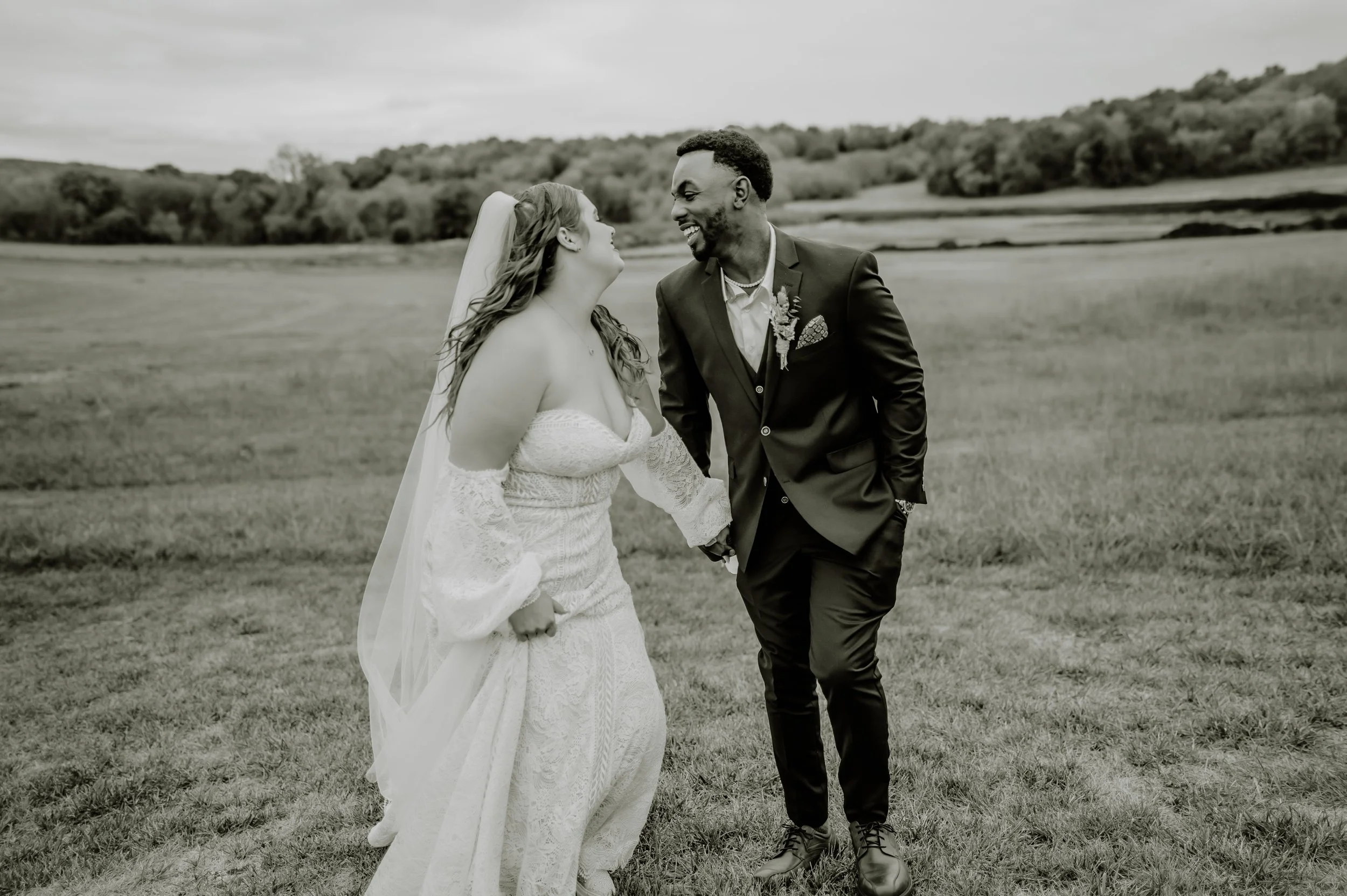 A bride and groom holding hands and smiling at each other outdoors in a grassy field with trees and hills in the background, with a cloudy sky.