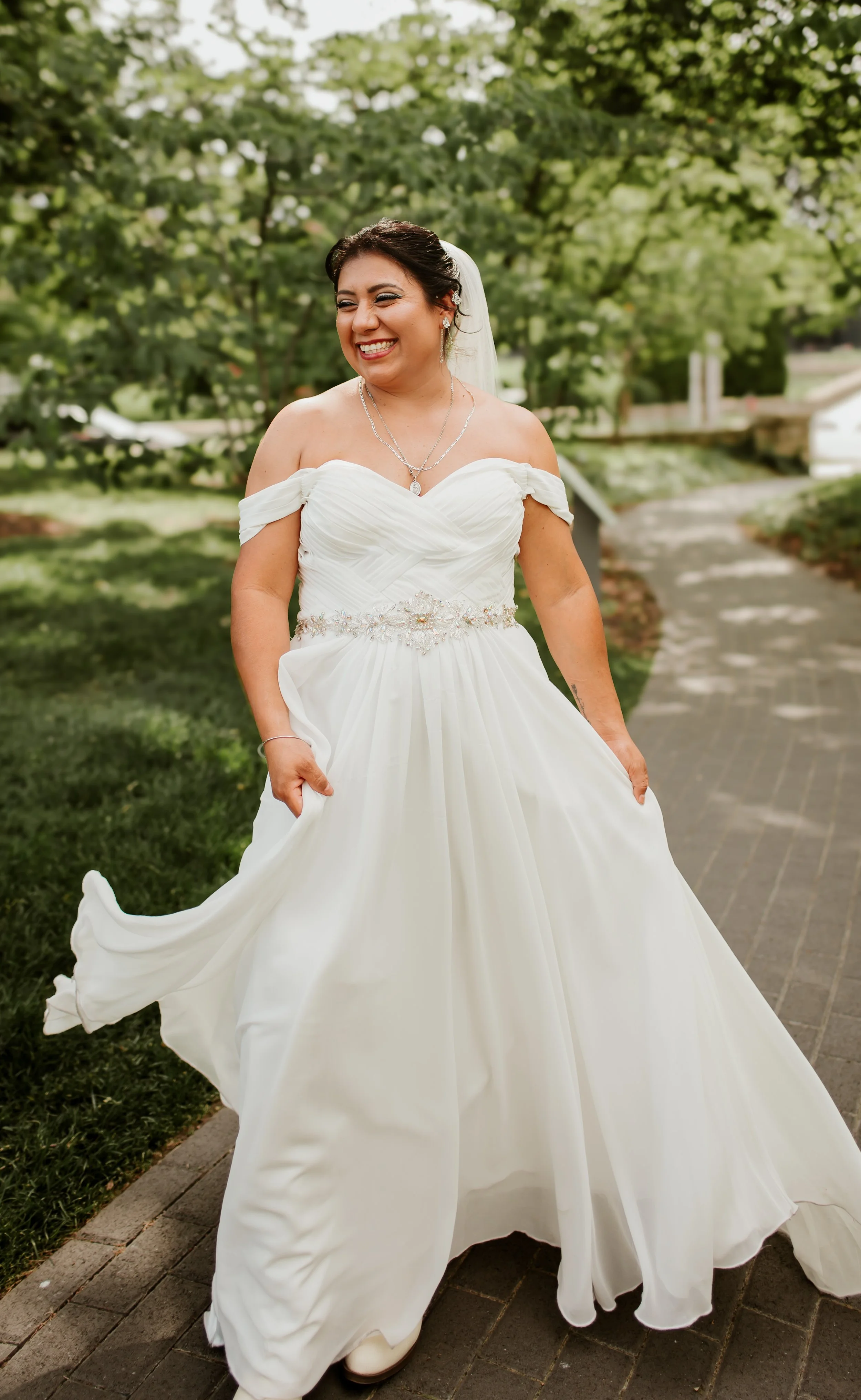 A smiling bride in a white off-shoulder wedding dress with lace and beading details, standing outdoors on a stone pathway with trees in the background.