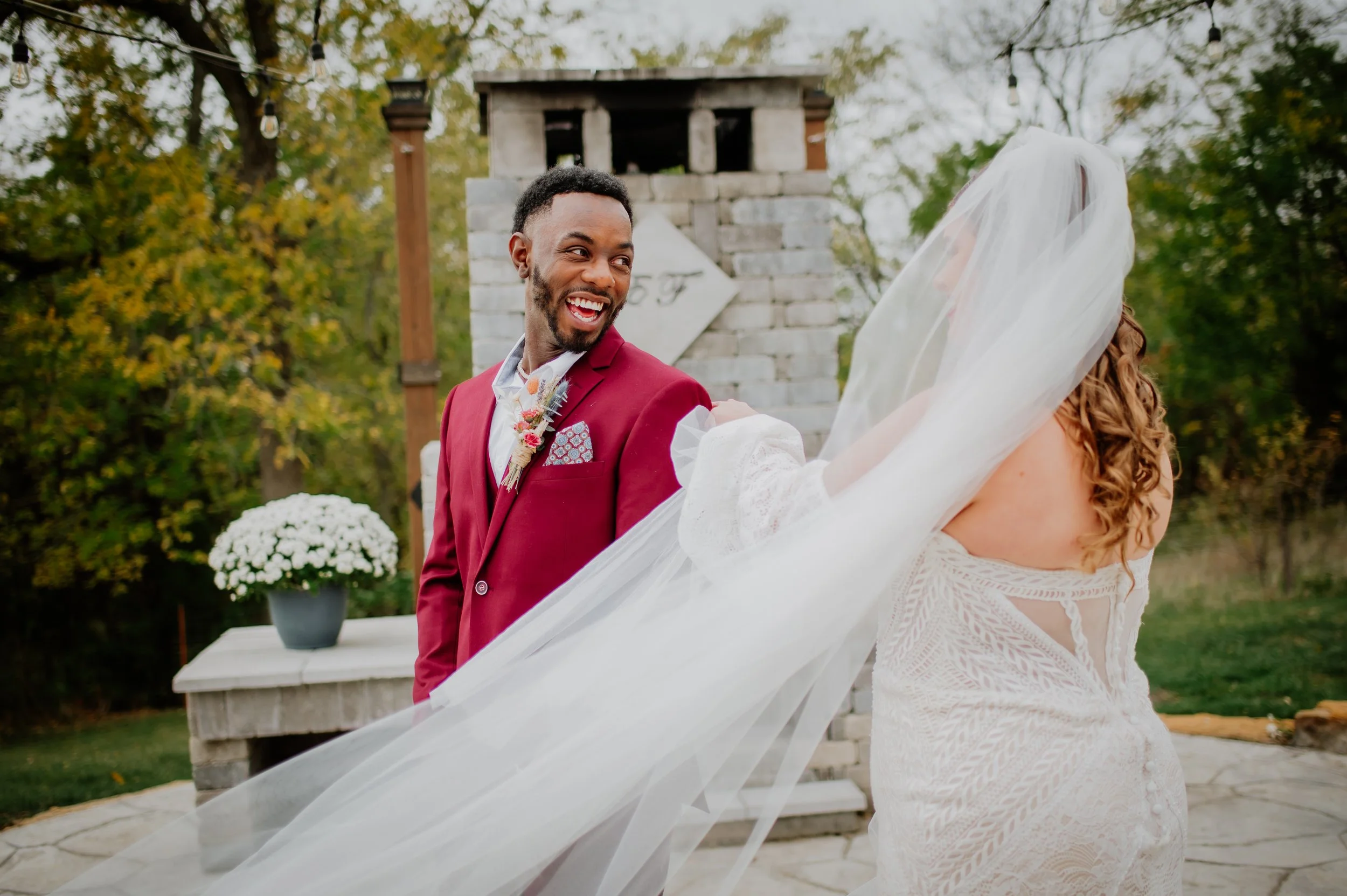 A smiling man in a red suit with a floral boutonniere stands outdoors next to a woman in a white lace wedding dress and veil, during a wedding ceremony, with trees, string lights, and a brick structure in the background.