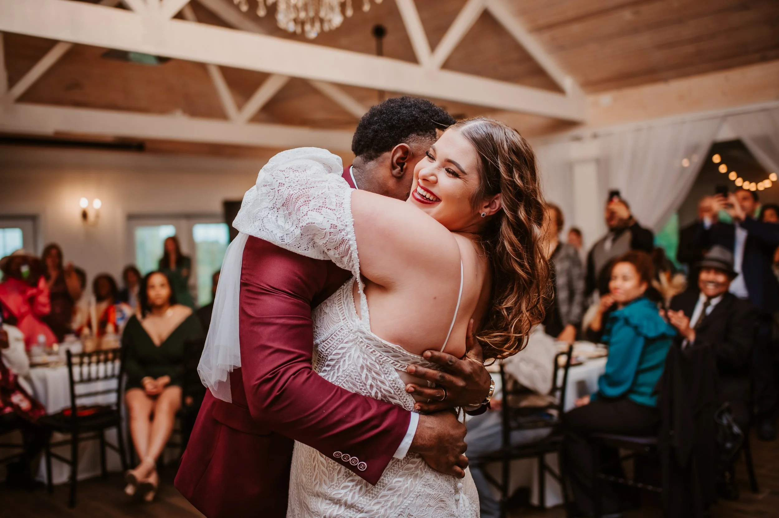 A couple hugging at a wedding reception, with the woman smiling and wearing a white lace dress, and the man in a burgundy suit. Guests are seated at tables in the background.