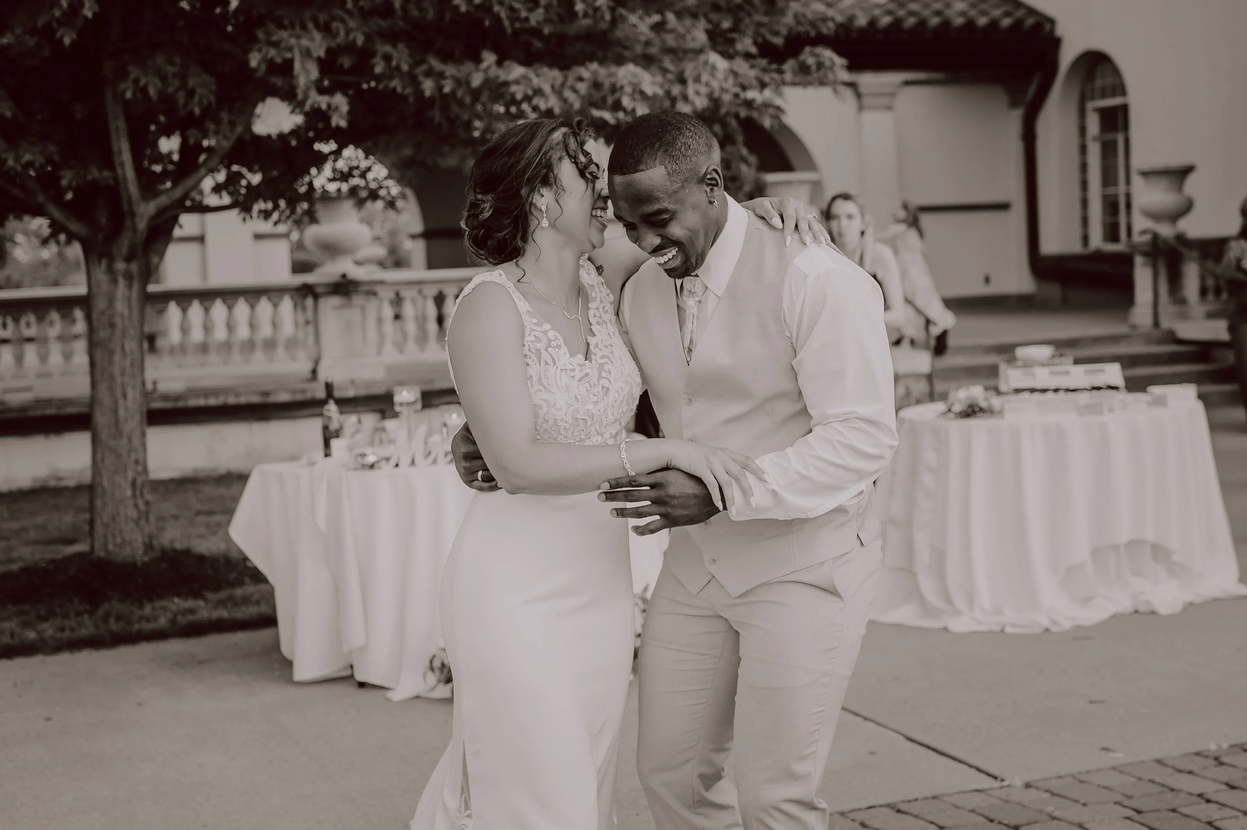 A couple is dancing outdoors at a wedding, smiling and embracing each other with friends and guests in the background.