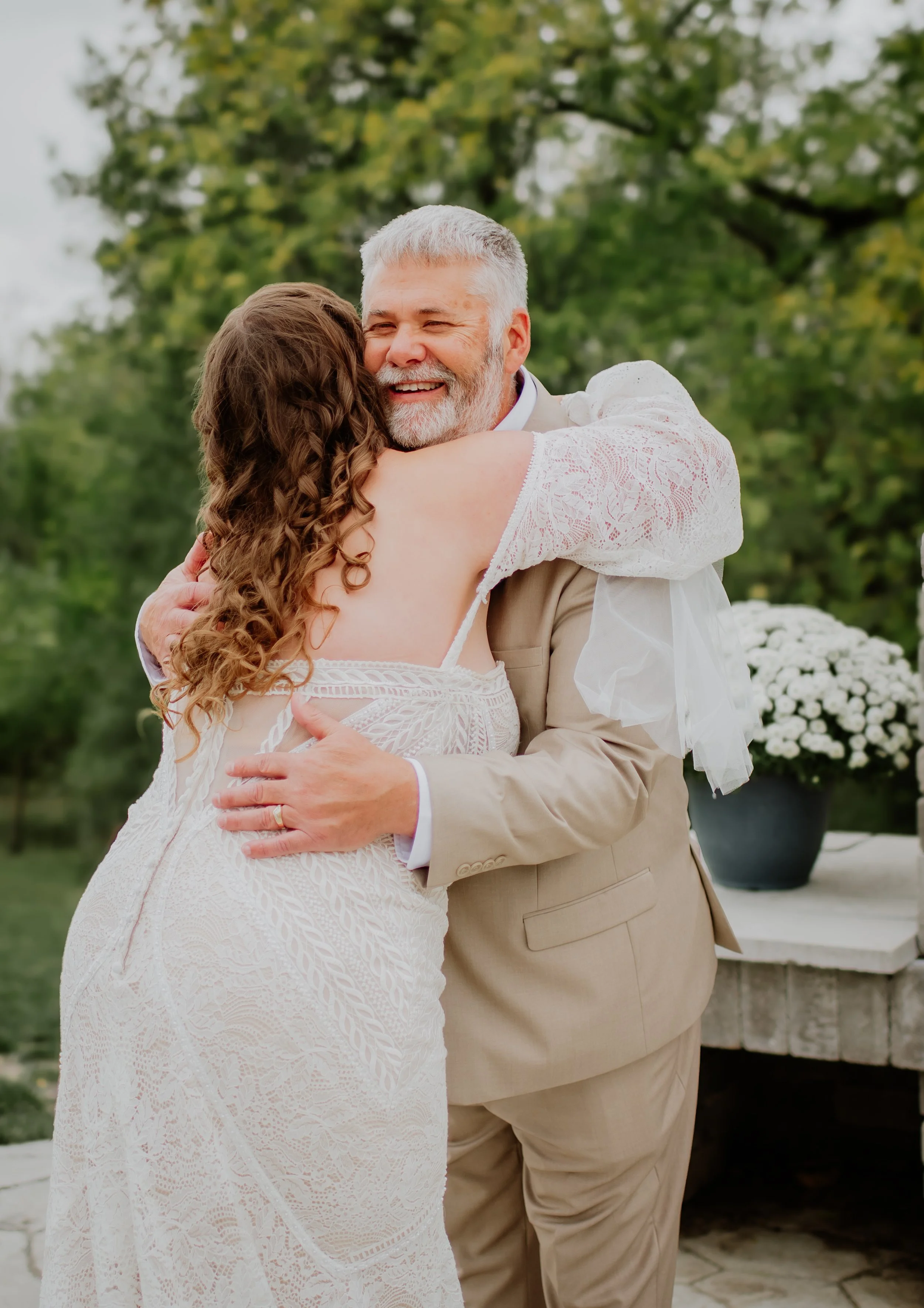 An older man with gray hair and a beard is hugging a woman in a white lace dress. They are outdoors, with greenery and a potted plant in the background, smiling and sharing a joyful moment.