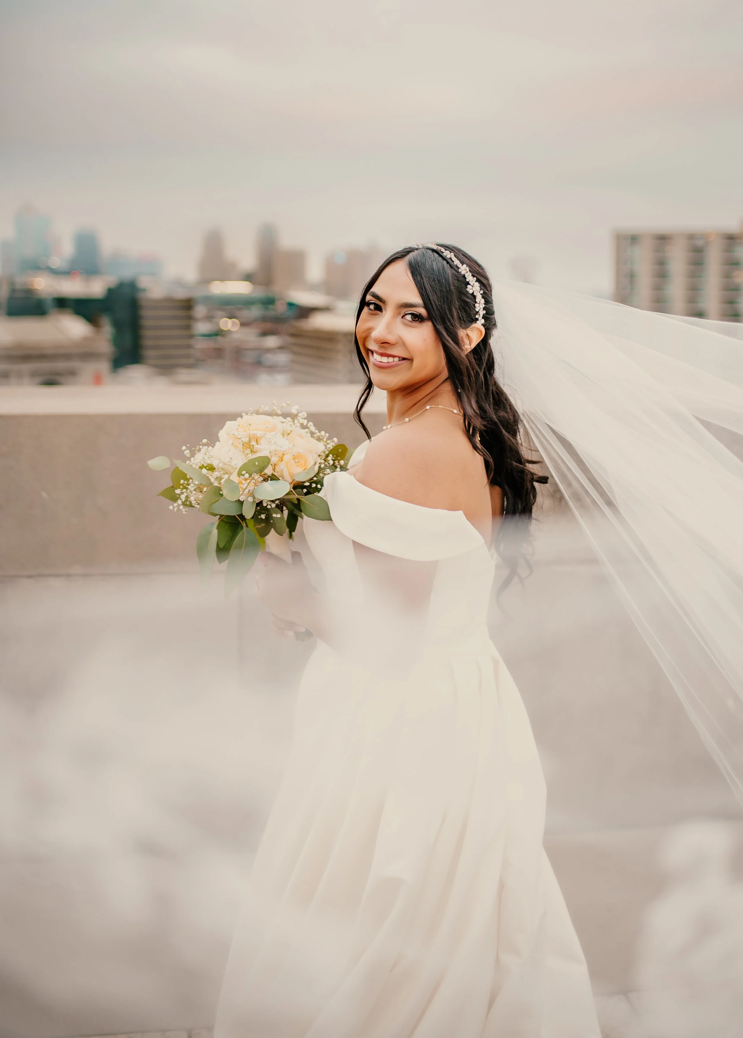 A smiling bride in a white off-shoulder wedding dress, holding a bouquet of cream roses and greenery, with a city skyline in the background.