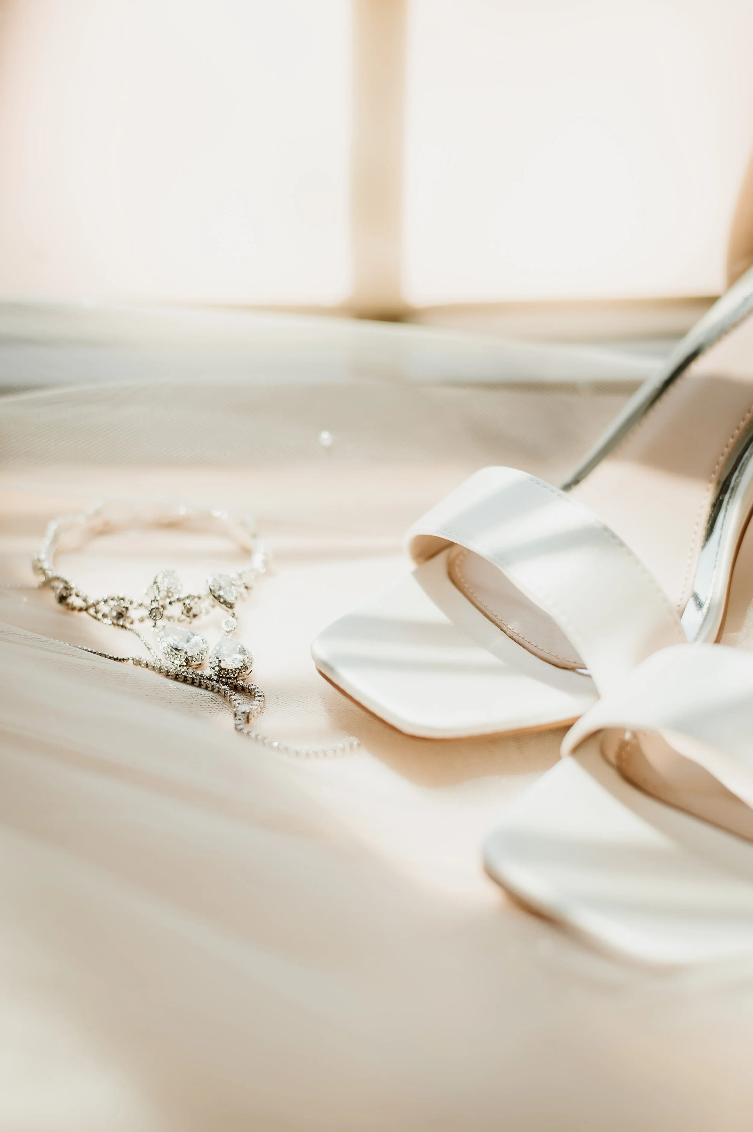 A pair of beige high-heeled shoes with white satin ribbons, a silver necklace with pendant, and a sparkly bracelet are arranged on a light-colored satin surface.