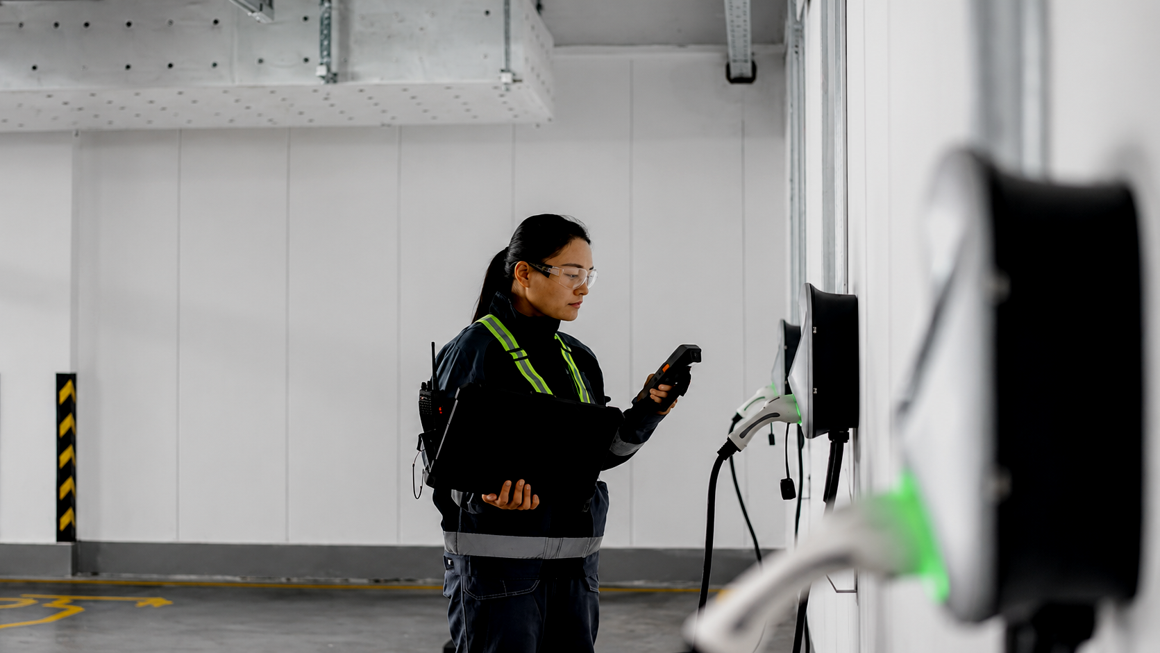 A female worker wearing safety glasses and a dark uniform stands in an industrial parking garage. She holds a clipboard in one hand and a device in the other, with electric vehicle charging stations on the wall in front of her.
