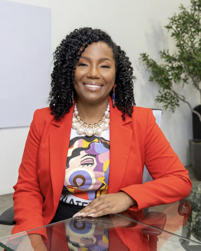 A woman sitting at a glass desk wearing a red blazer and colorful graphic t-shirt, with curly black hair, pearl necklace, and earrings, smiling in an office setting.