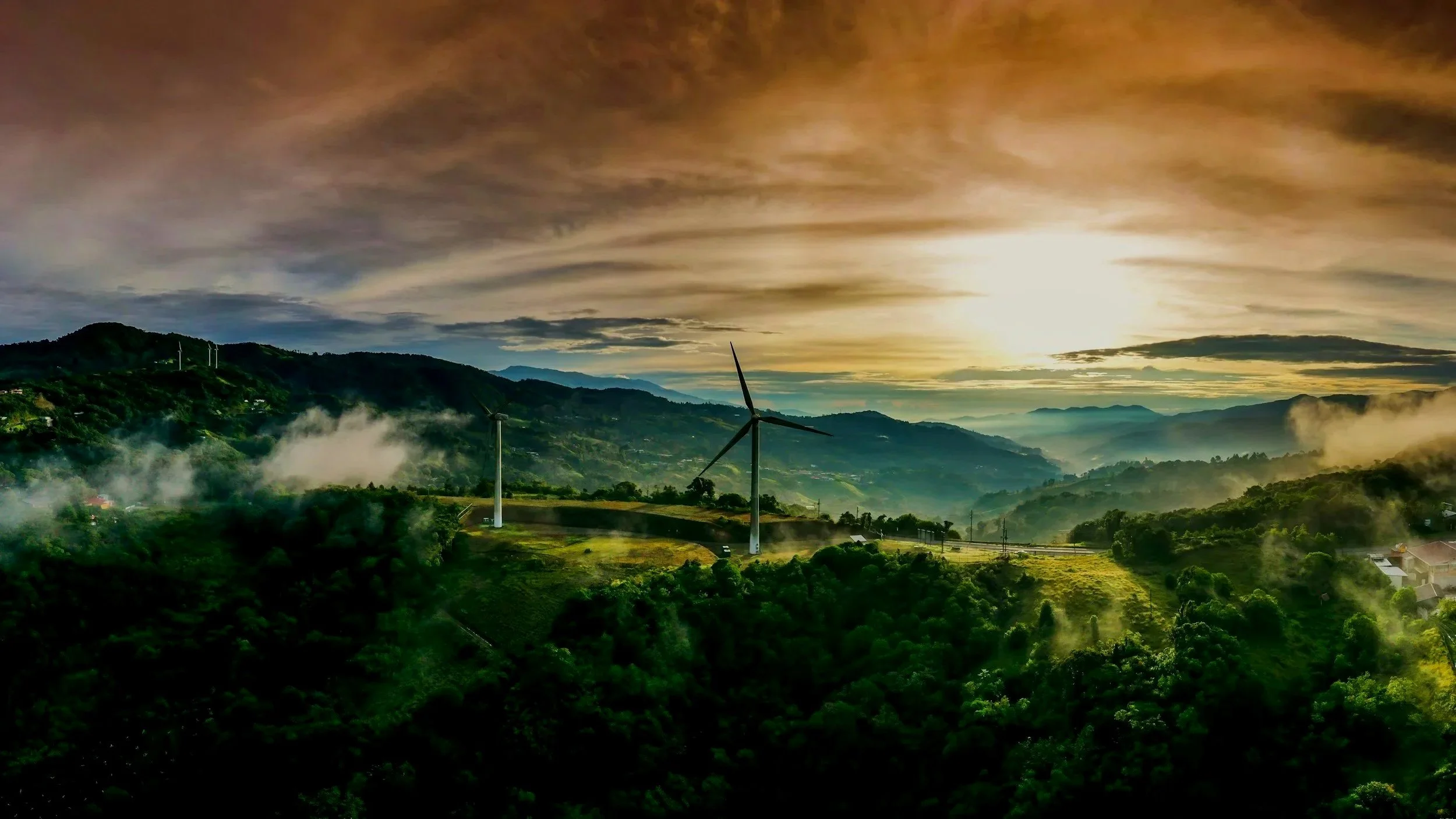 A scenic landscape featuring rolling hills covered in lush green vegetation, wind turbines scattered across the hills, mist rising from the valley, and a dramatic sky with golden hues during either sunrise or sunset.