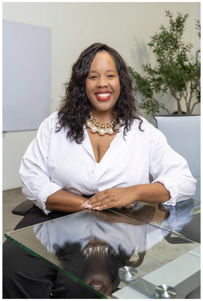 A woman with curly hair and a pearl necklace sits at a glass table, smiling at the camera in an office setting.