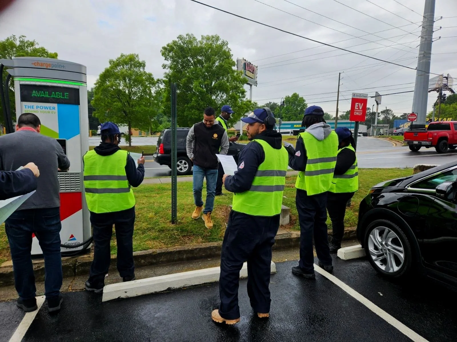 Group of people, some wearing yellow safety vests, gathered near electric vehicle charging station on wet pavement on overcast day, with street and trees in background.