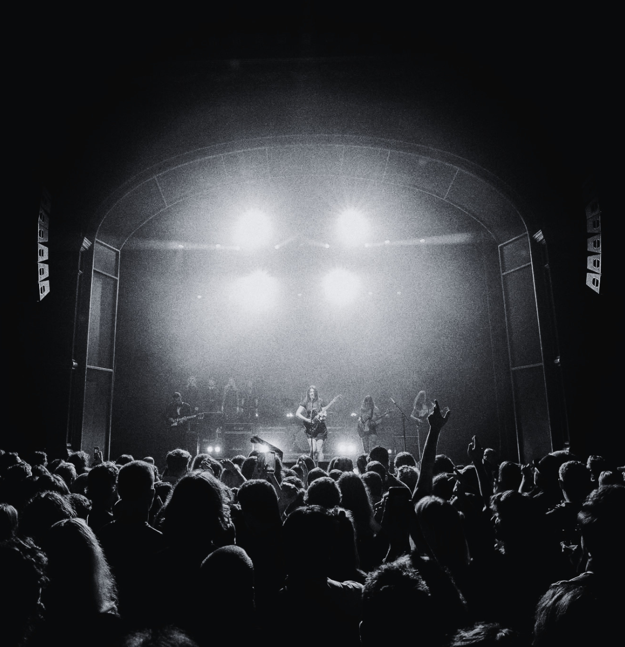 Black and white photo of a live concert with a band playing on stage under bright lights and a crowd watching, some with hands raised.