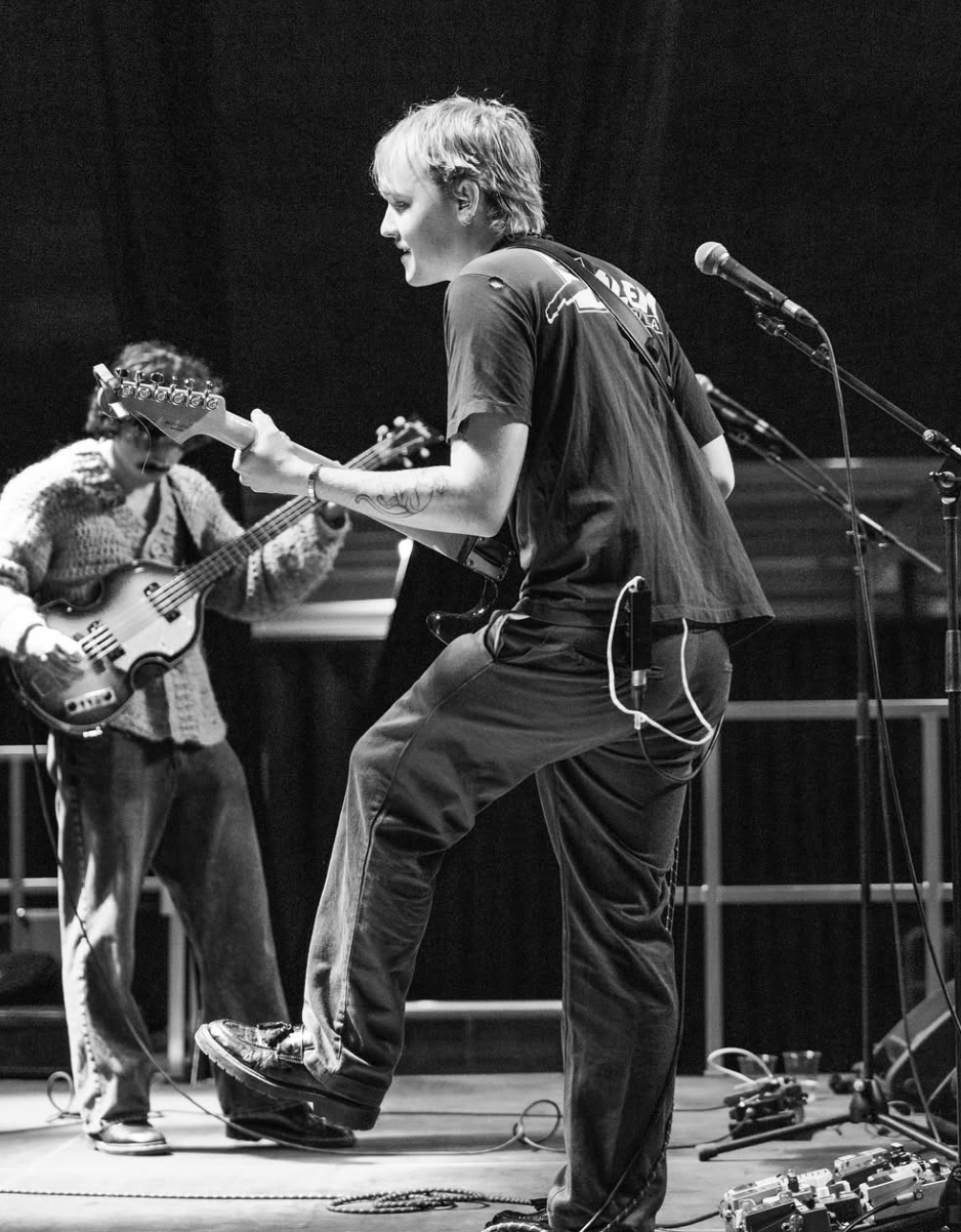 Two musicians performing on stage, one playing an electric guitar and the other playing a bass guitar, with musical equipment around them, in a black-and-white setting.