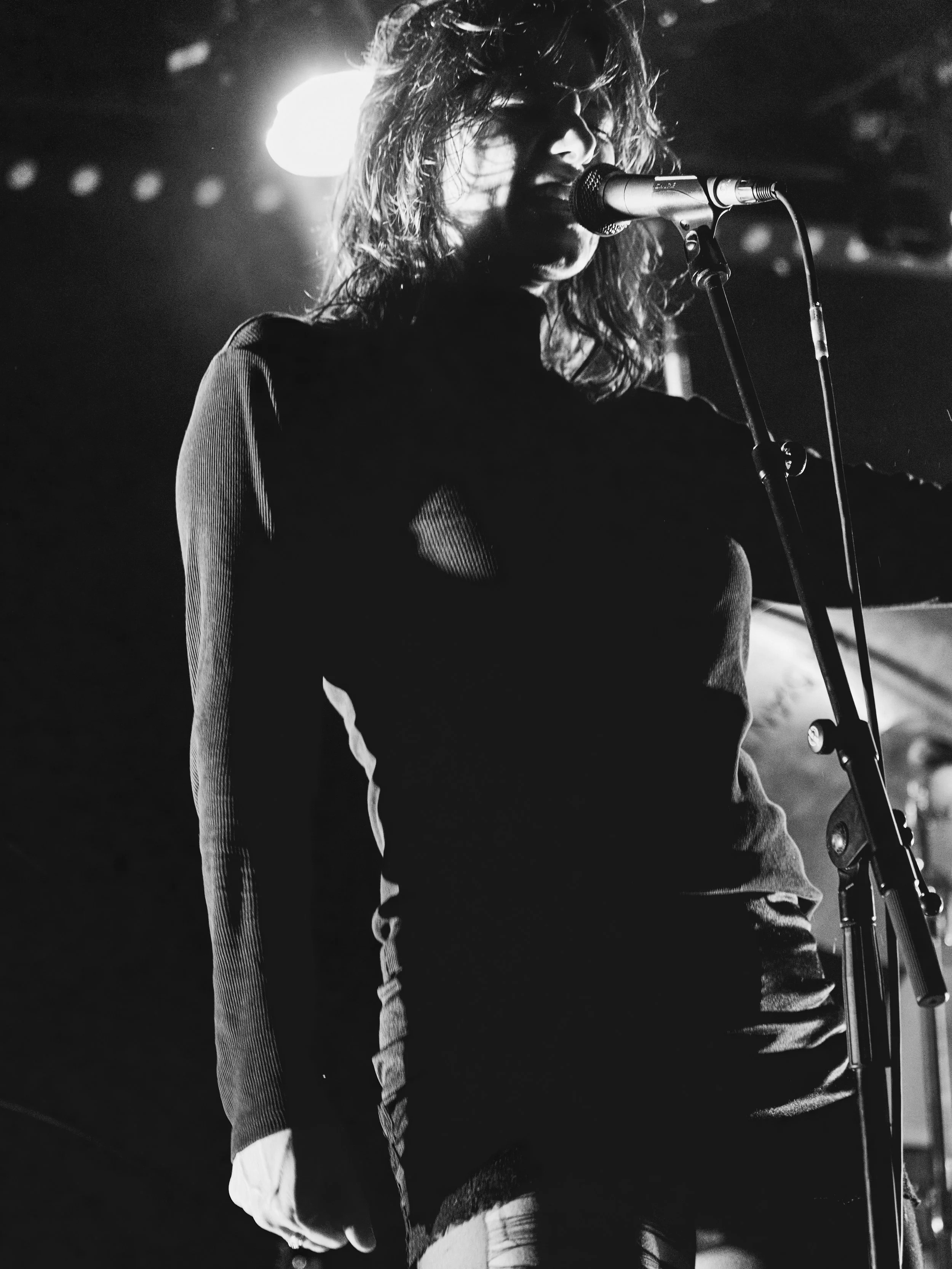 Black and white photo of a person performing on stage, singing into a microphone with closed eyes and expressive facial expression, backlit with bright light.