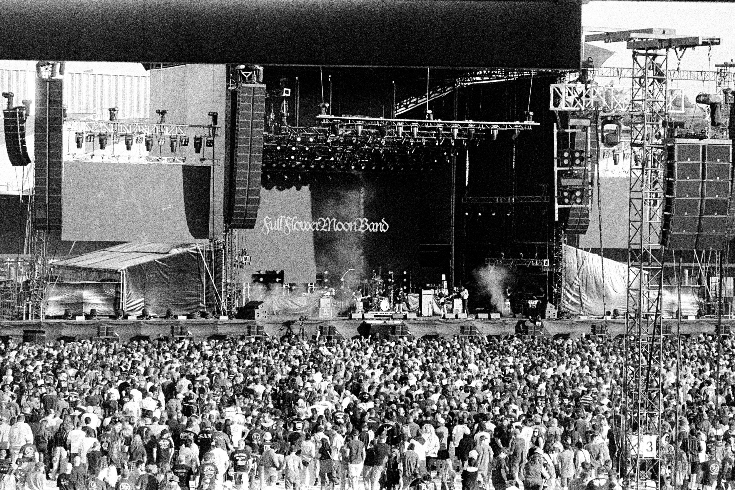 A black and white photo of a large outdoor concert stage with a crowd of people in front. The stage has various lighting and sound equipment, with a band performing and a large screen displaying the band's name, 'Full Flower Moon Band'.