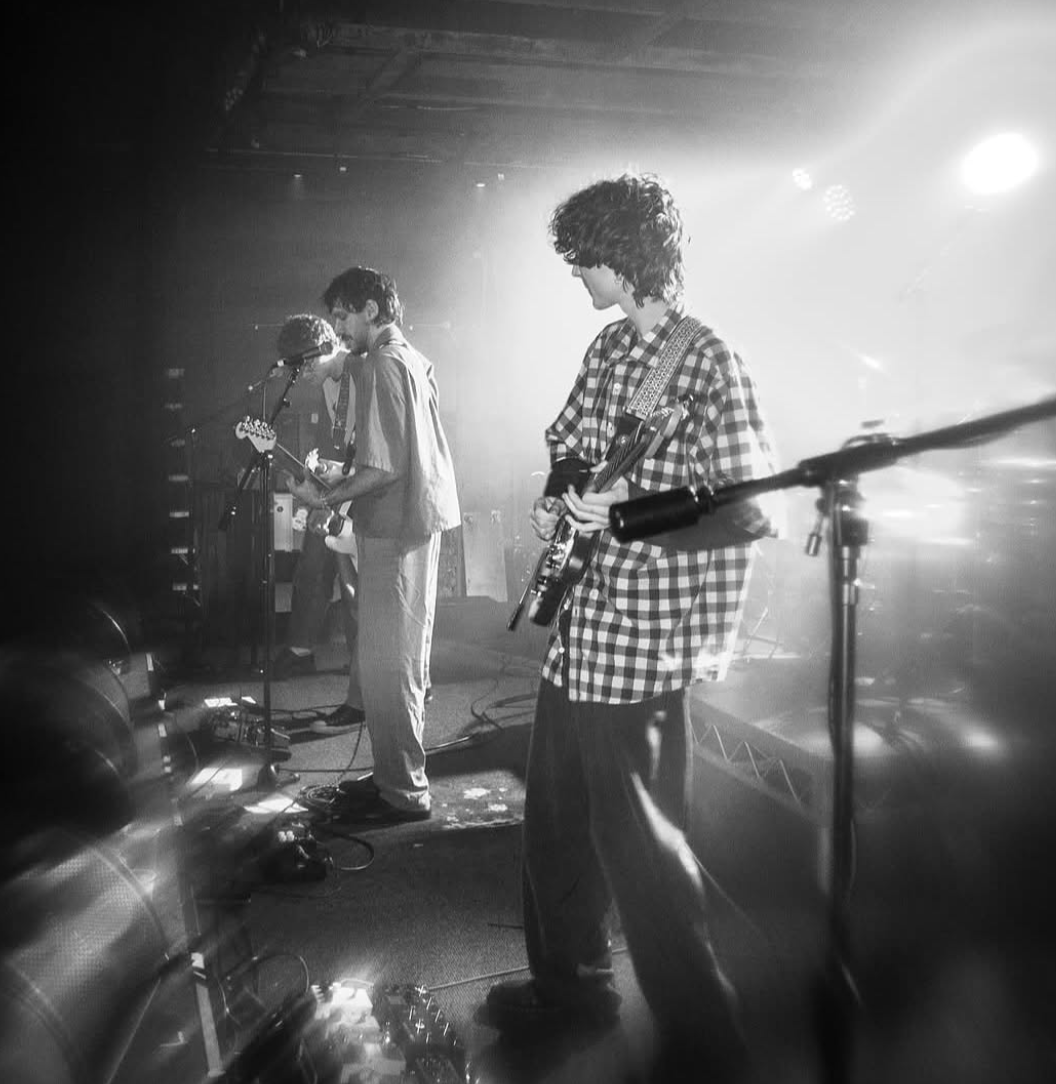 Black and white photo of a band performing on stage with three musicians playing guitars and singing into microphones, stage lights shining from behind.