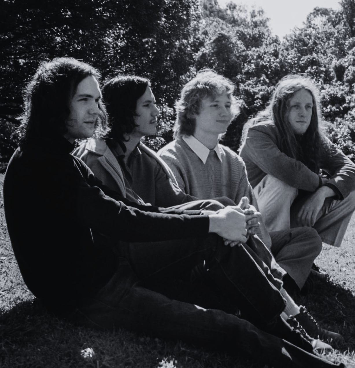 Four young men sitting outdoors on grass with trees in the background, looking to the right. Black and white photograph.