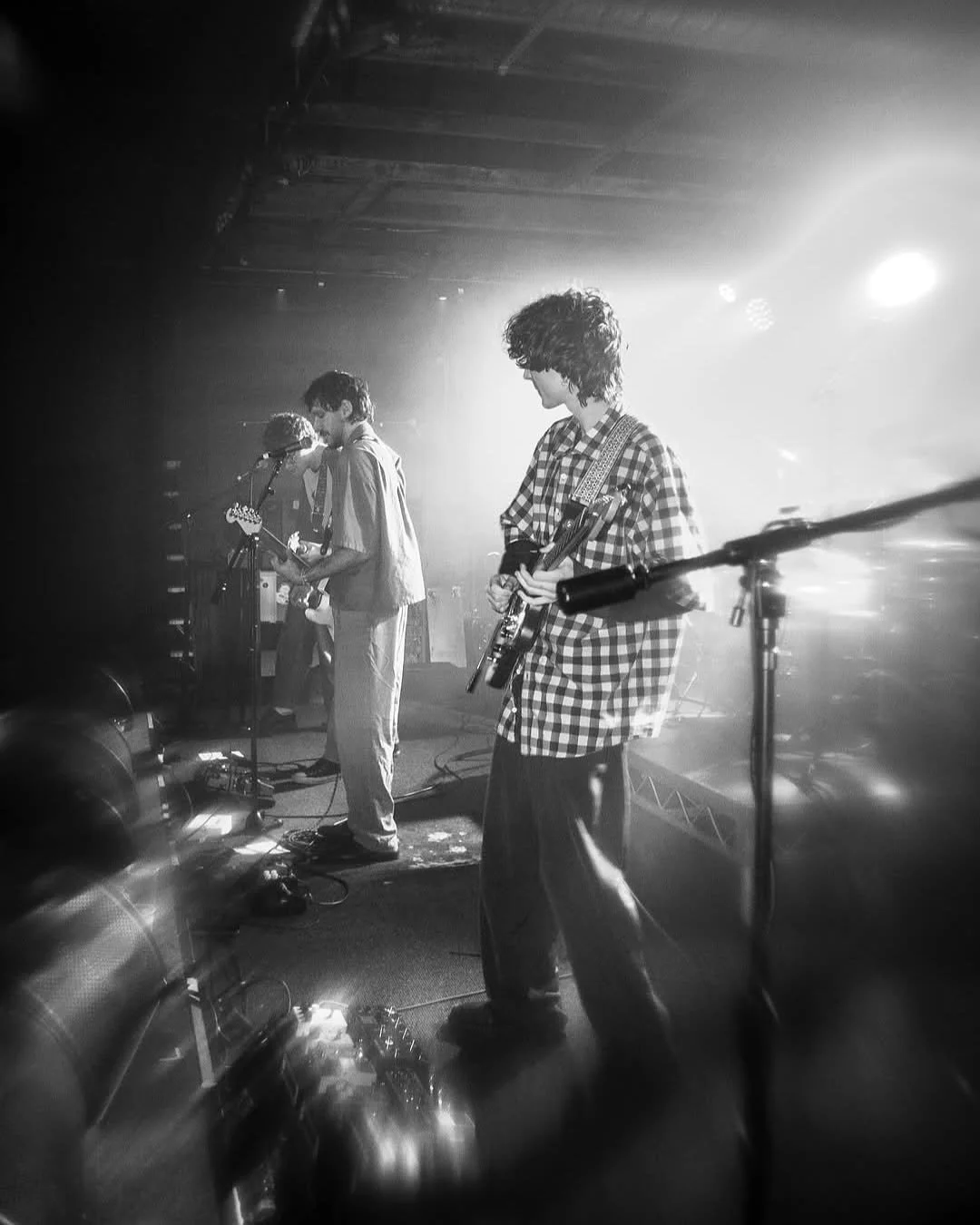Black and white photo of a band performing on stage with three musicians playing guitars and singing, illuminated by stage lights.