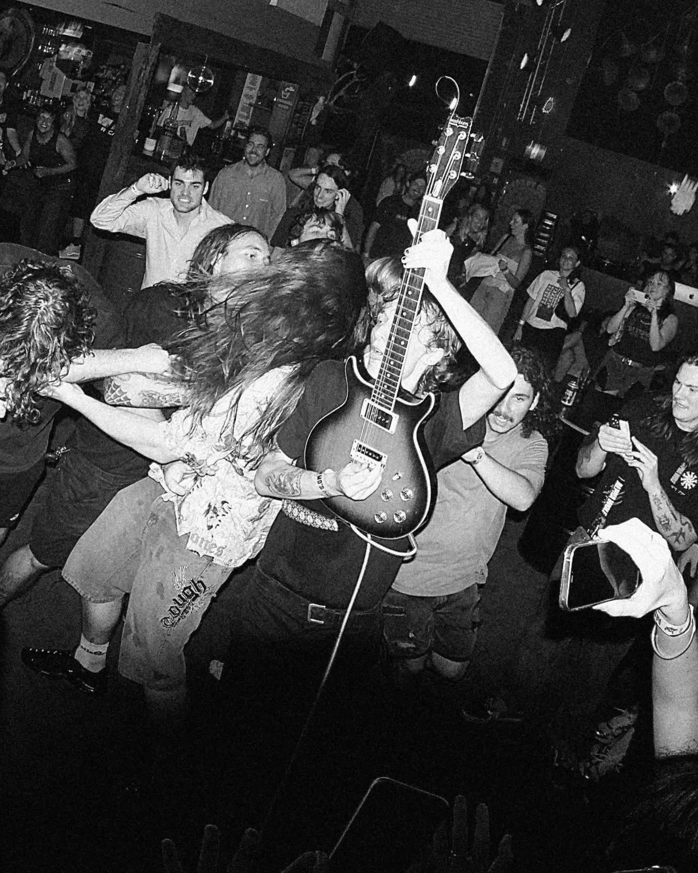 A lively social scene at a bar or club with people enjoying music, singing, dancing, and taking photos, in black and white.