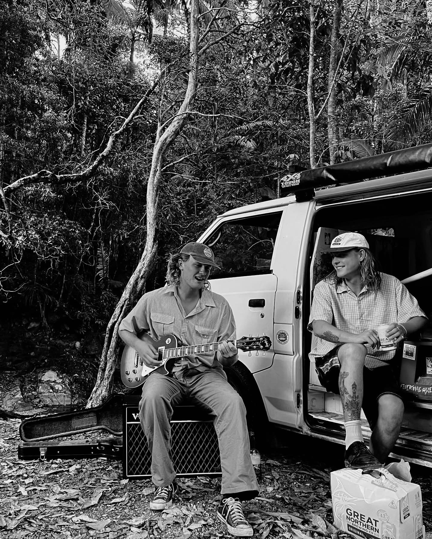 Two women sitting outdoors in front of a white vehicle in a wooded area. One woman is playing an electric guitar, wearing a baseball cap and casual clothes, sitting on a small crate with an amplifier nearby. The other woman, with tattoos and wearing a baseball cap, is sitting on the vehicle's step, holding a drink and smiling. There are miscellaneous items on the ground, including a paper bag labeled 'GREAT NORTHWEST BREWING'.