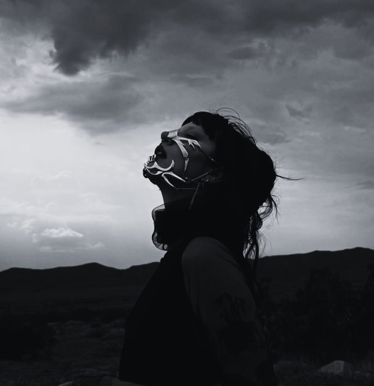 A person wearing a mask with intricate designs, standing outdoors against a cloudy sky and mountain landscape, in black and white.