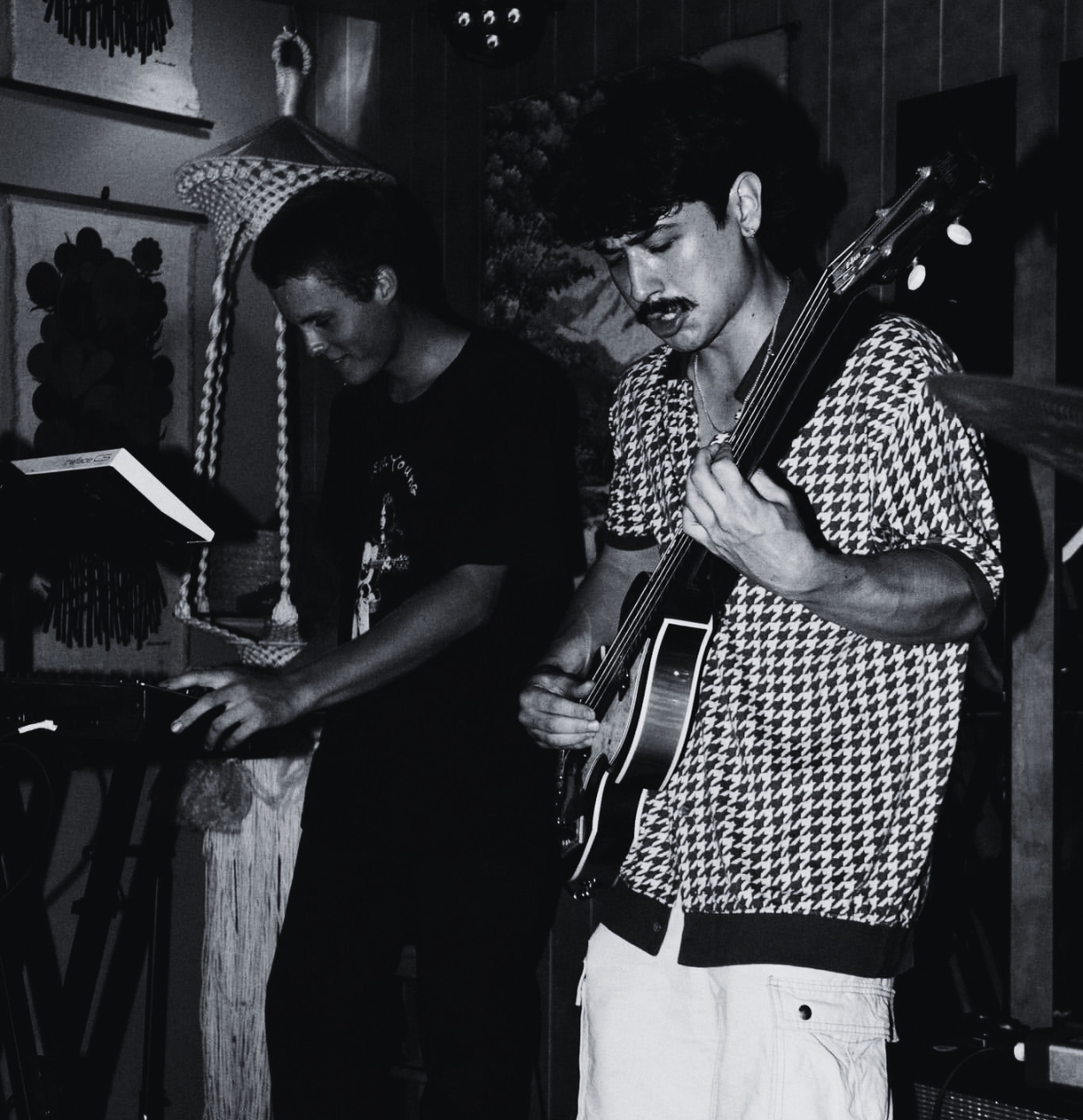 Two musicians playing instruments, one on a keyboard and the other on an acoustic guitar, in a cozy indoor setting with wall art and decorative items in the background.