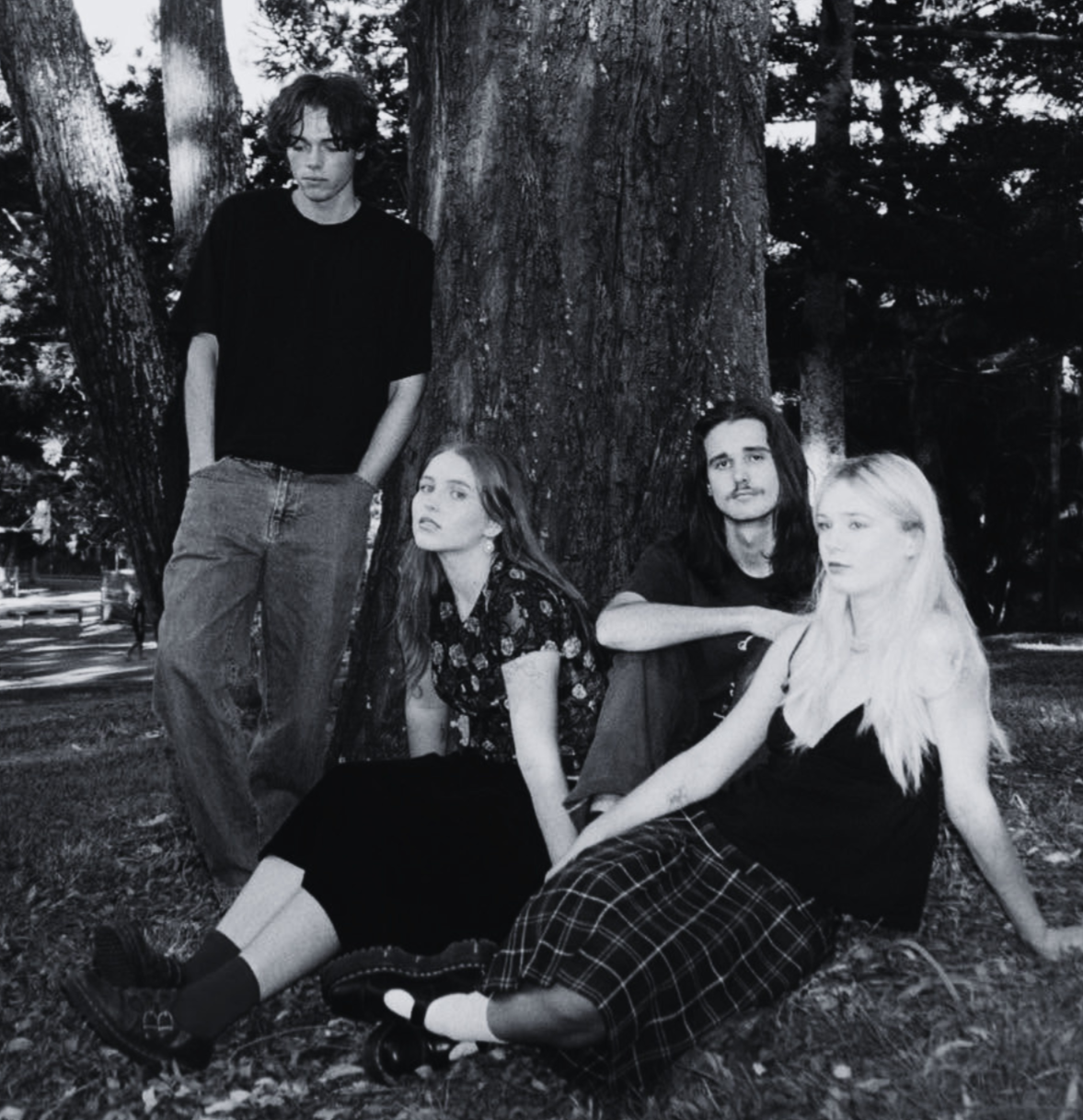 Four young people in a park, sitting and standing around a large tree, with leaves and other trees visible in the background. The image is black and white.
