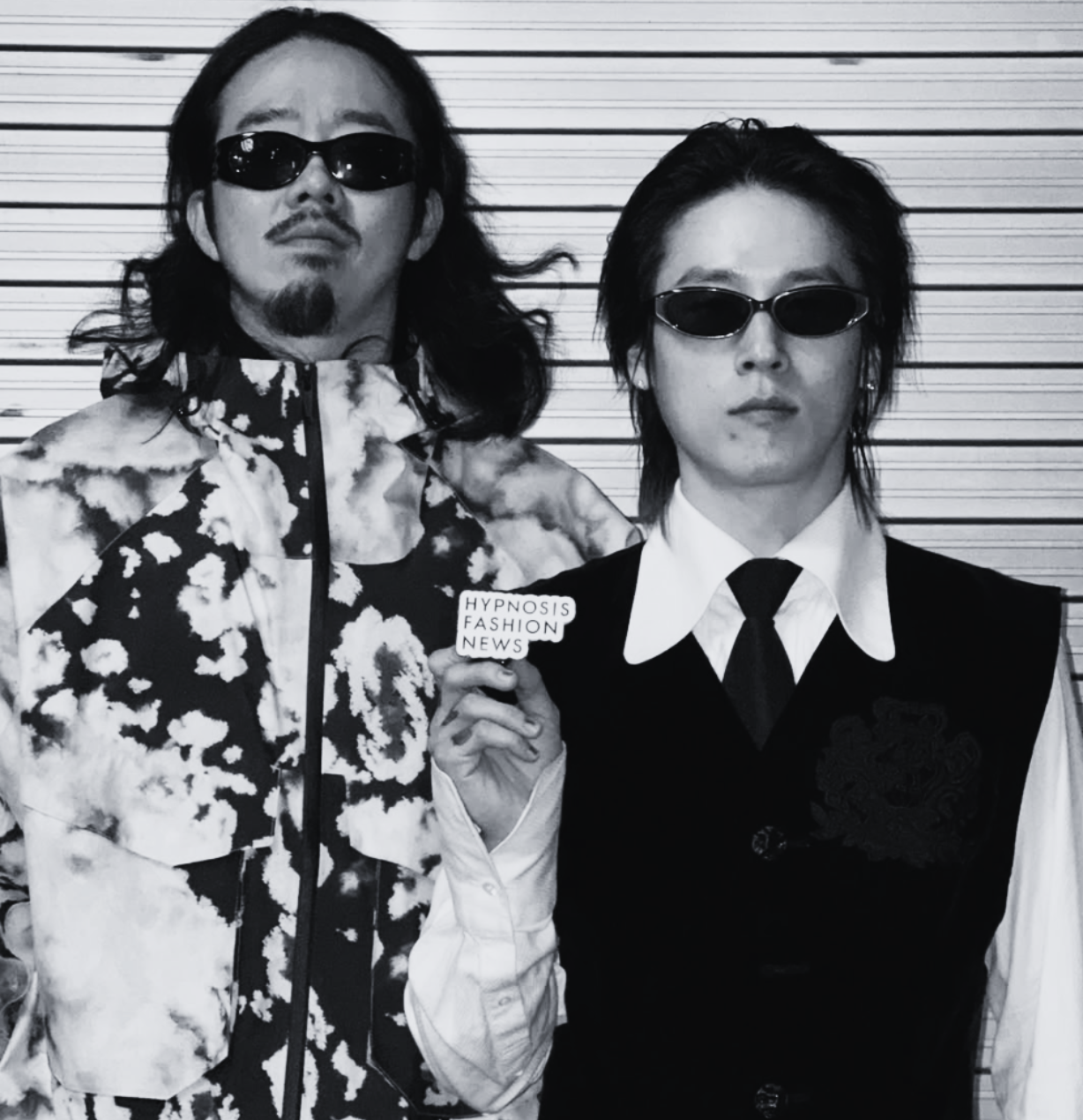 Two young men with long hair and sunglasses standing side by side in front of a horizontal striped background. One is wearing a patterned jacket and holding a sign that says "HYPNOSIS FASHION NEWS."