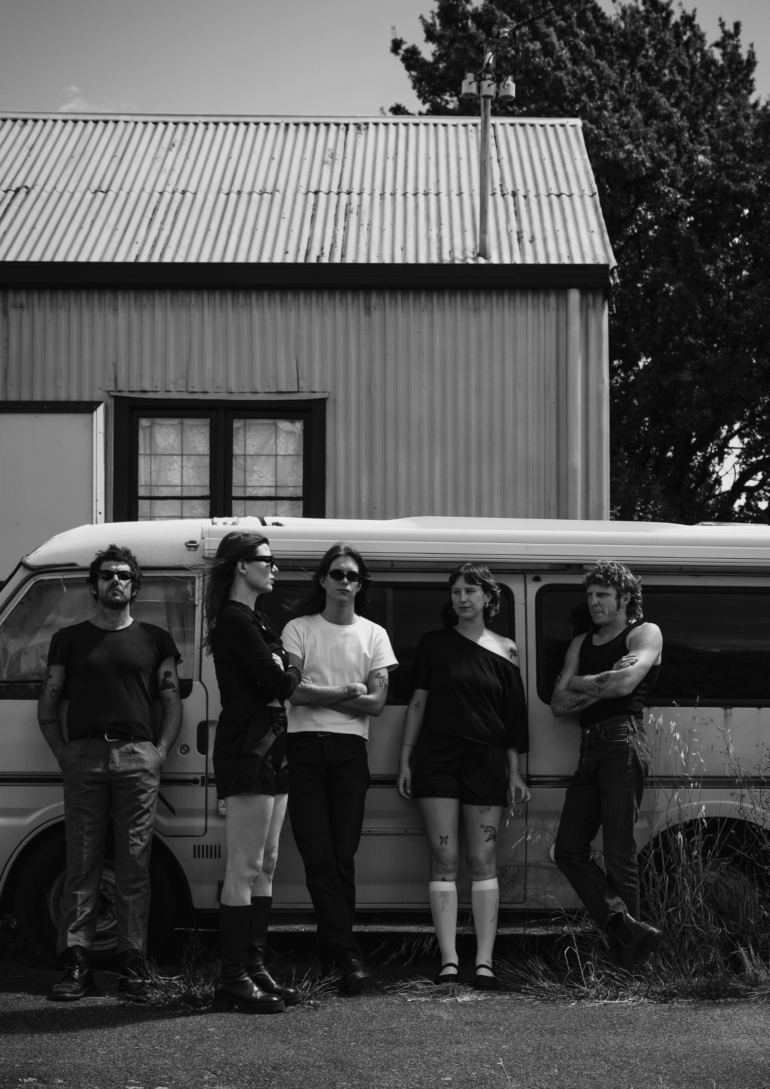 A group of five young adults standing in front of a van, with a building and a tree in the background, in black and white.
