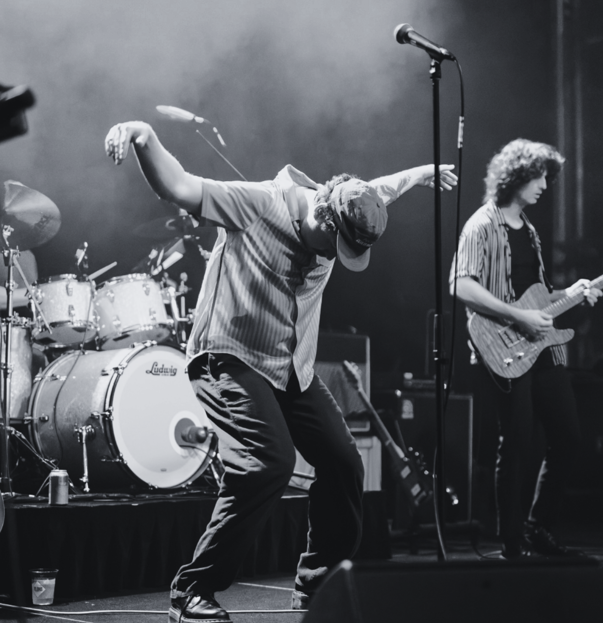A musician dancing on stage with a hat, a striped shirt, and baggy pants, with a band playing instruments in the background, including a drummer and a guitarist, in a black-and-white concert scene.