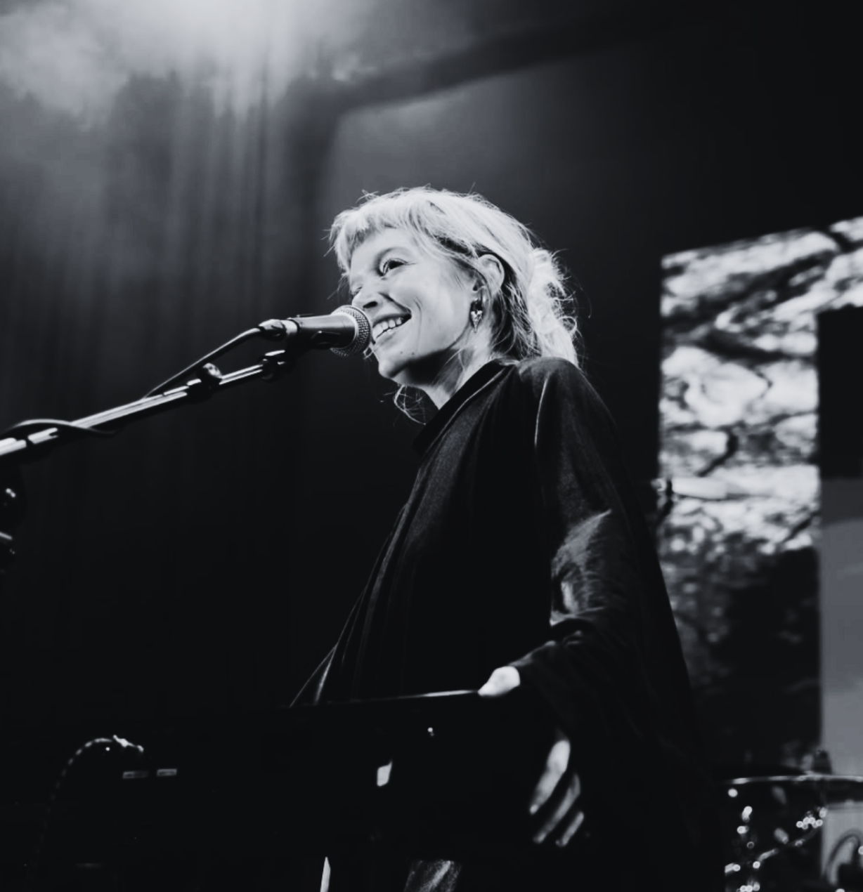 Black and white photo of a woman singing into a microphone and playing a keyboard on stage, smiling, with a textured background