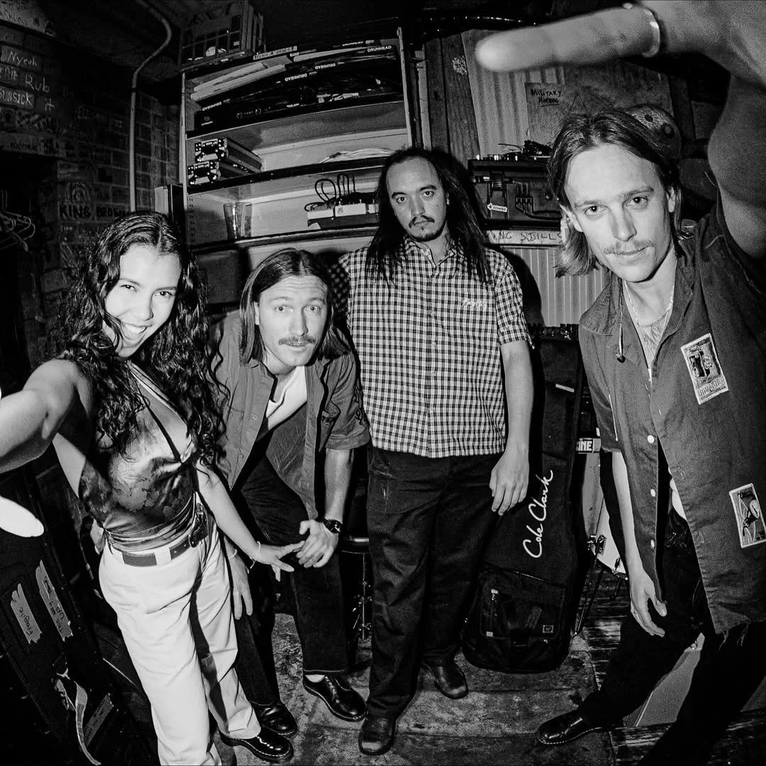 Black and white photo of four people in a music practice room, with musical equipment in the background. Two men are on the right, and a woman and man are on the left, all looking at the camera.