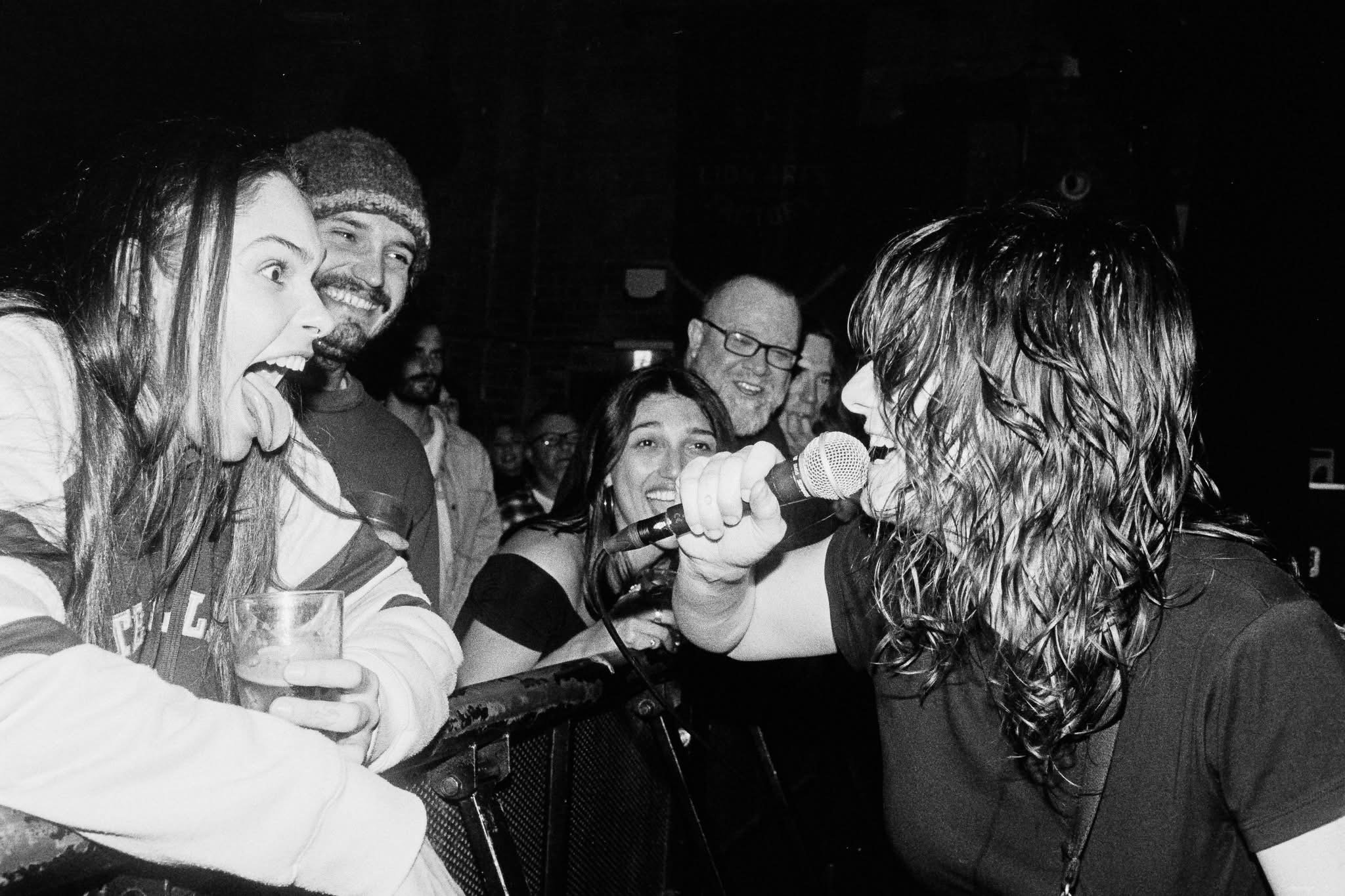 Group of people at a social event, with one woman with curly hair holding a microphone, others smiling and engaging in conversation, in a dimly lit setting.