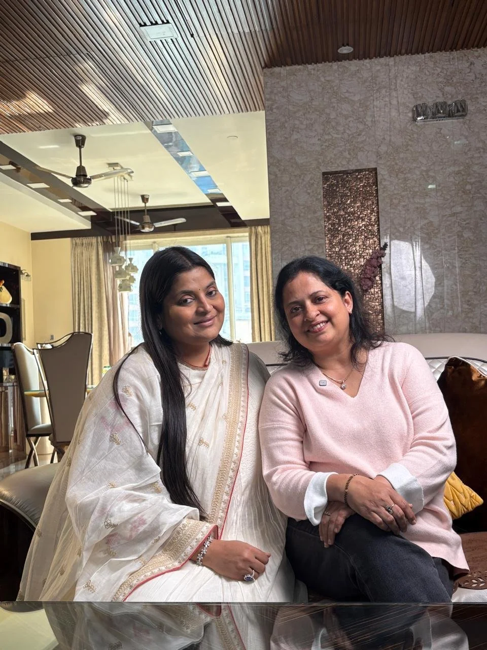Two women sitting on a beige sofa in a modern living room, smiling at the camera. One woman is dressed in traditional white attire, and the other is wearing a light pink top.