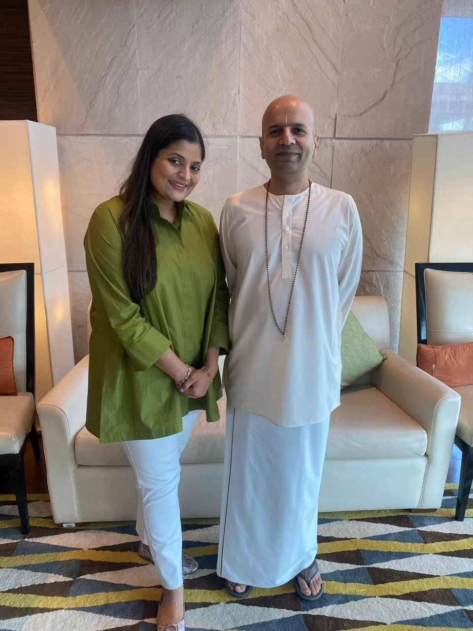 A woman in a green tunic and white pants standing next to a man in traditional white Indian attire with a mala necklace, in a modern indoor setting.