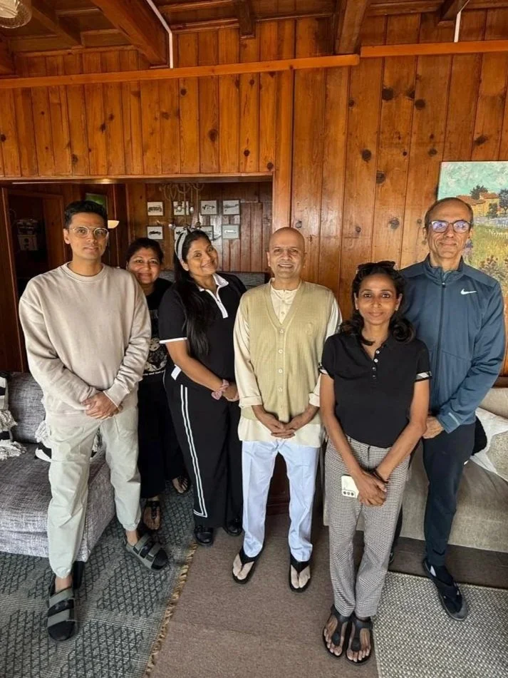 Six people standing indoors in front of wooden wall, smiling at the camera, dressed casually.