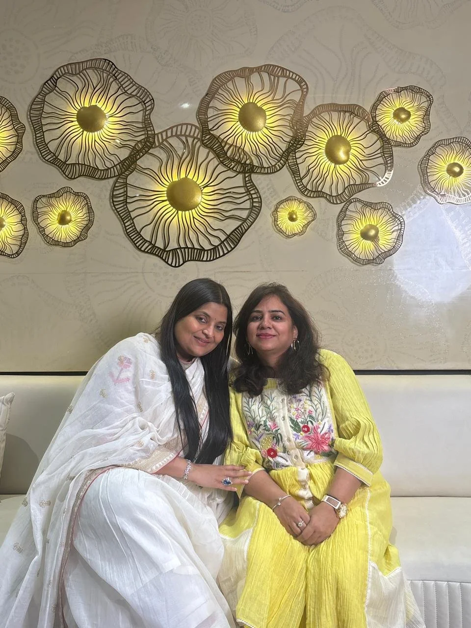 Two women sitting on a cream-colored bench in front of a decorative wall with yellow flower-shaped lights. One woman wears a white traditional dress, and the other wears a yellow dress with floral embroidery.