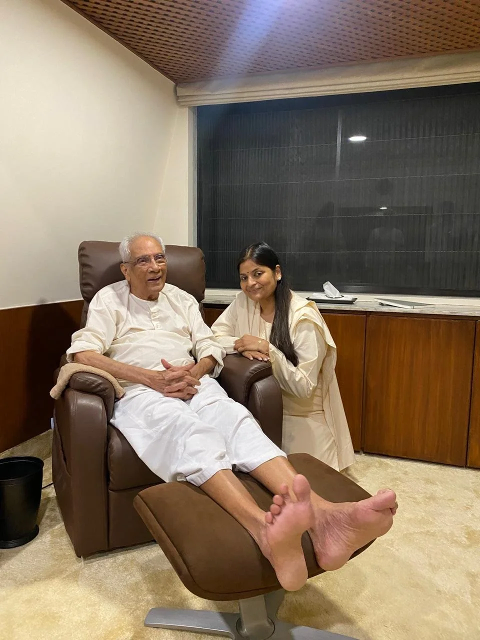 Older man sitting in a brown armchair with his feet up on a footrest, wearing white traditional clothing, smiling, with a woman kneeling beside him, resting her hand on his arm, in a room with wooden paneling, a large window, and a beige carpet.