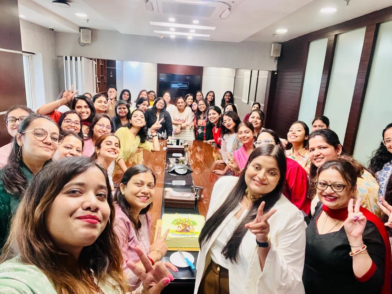 Group of women gathering around a conference table, smiling and making peace signs, celebrating with a cake, in a modern conference room.