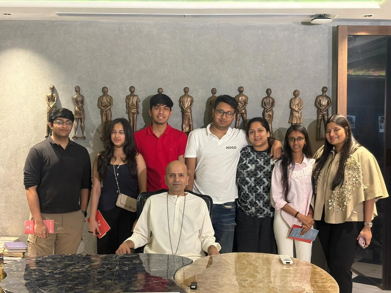 Group of eight people, including one seated man and seven standing, posing indoors in front of a wall with sculptures of people. The group is diverse in appearance and attire, with some holding books or pamphlets.