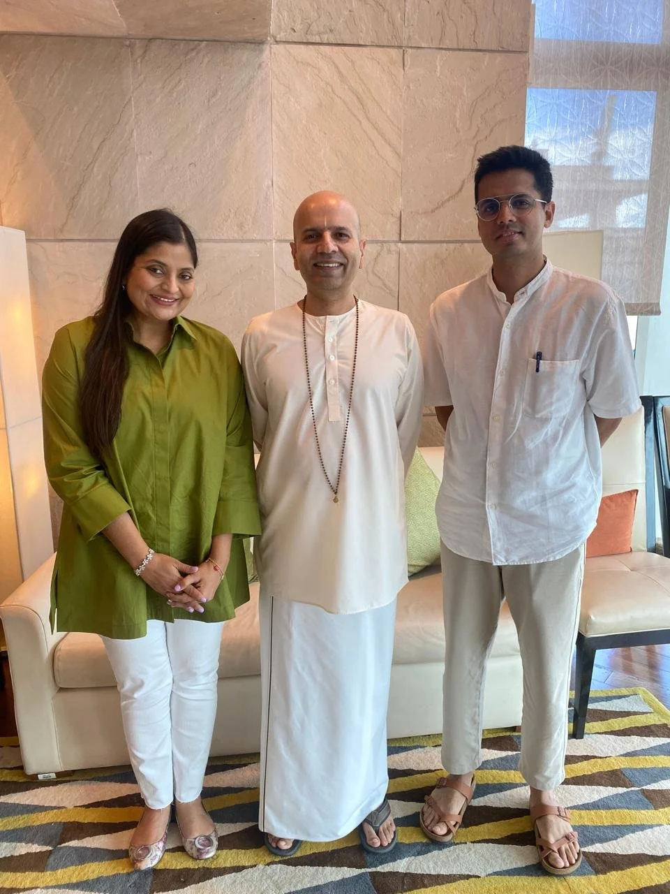Three people standing side by side indoors, smiling at the camera. Woman on the left wearing a green top and white pants, man in the center wearing traditional white Indian attire with a prayer bead necklace, man on the right wearing a white shirt, b