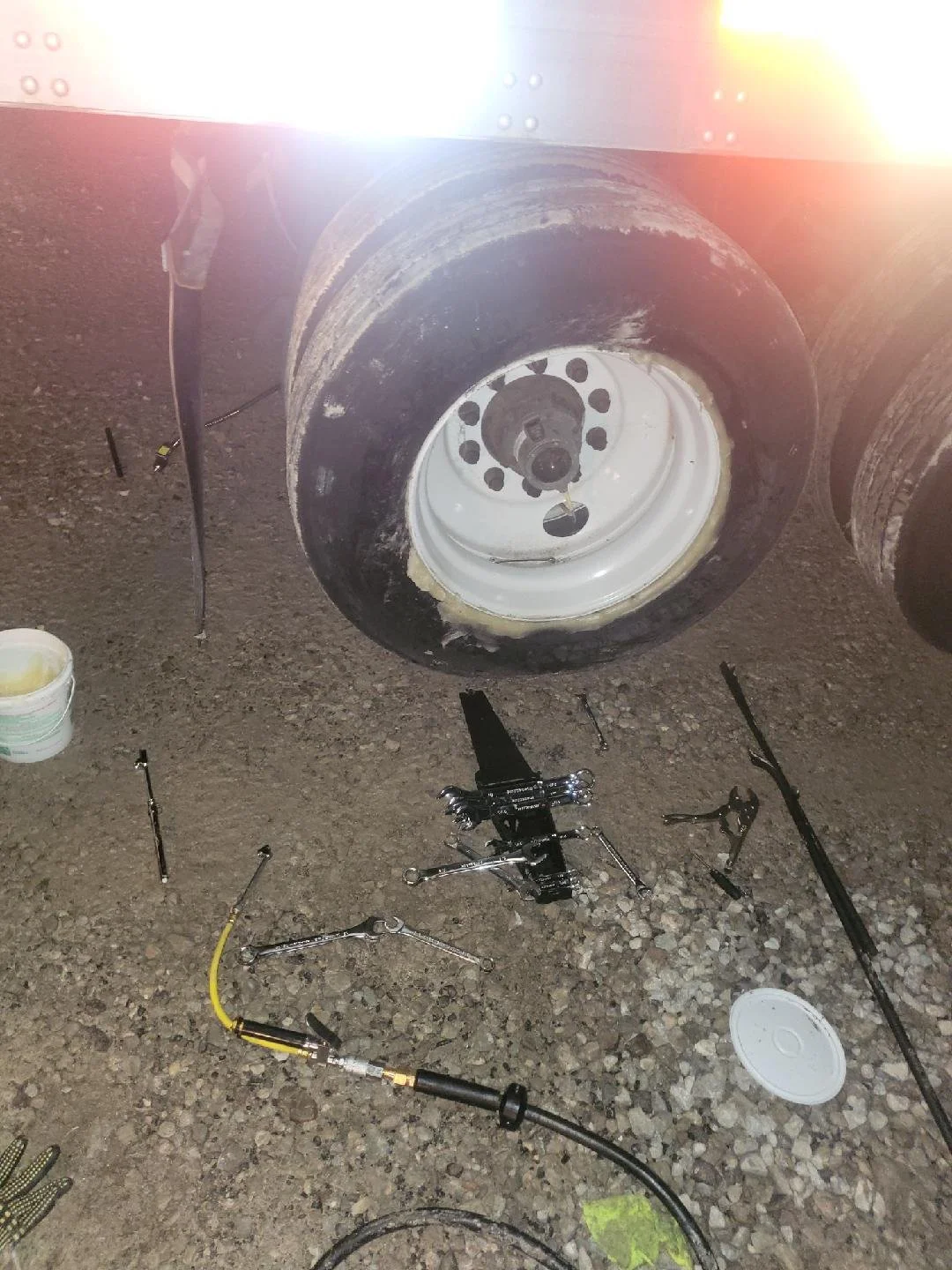 Close-up of a large truck's wheel, with the tire removed, surrounded by various tools and equipment on the ground, including a hydraulic jack, ratchets, and a bucket.