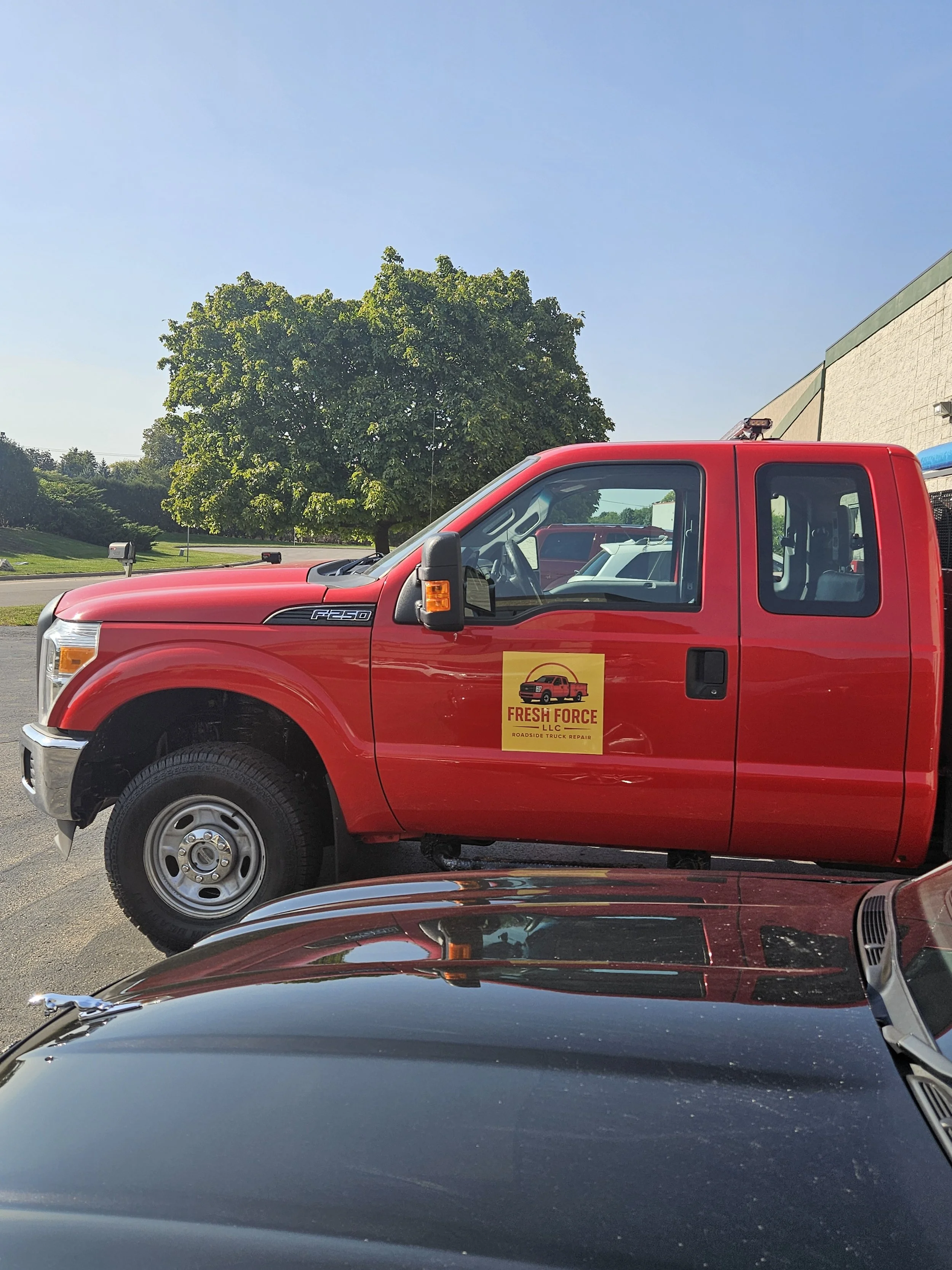 Red roadside truck with a yellow sign that says "Fresh Force LLC Roadside Truck Repair" parked beside a building, with trees and a clear blue sky in the background.