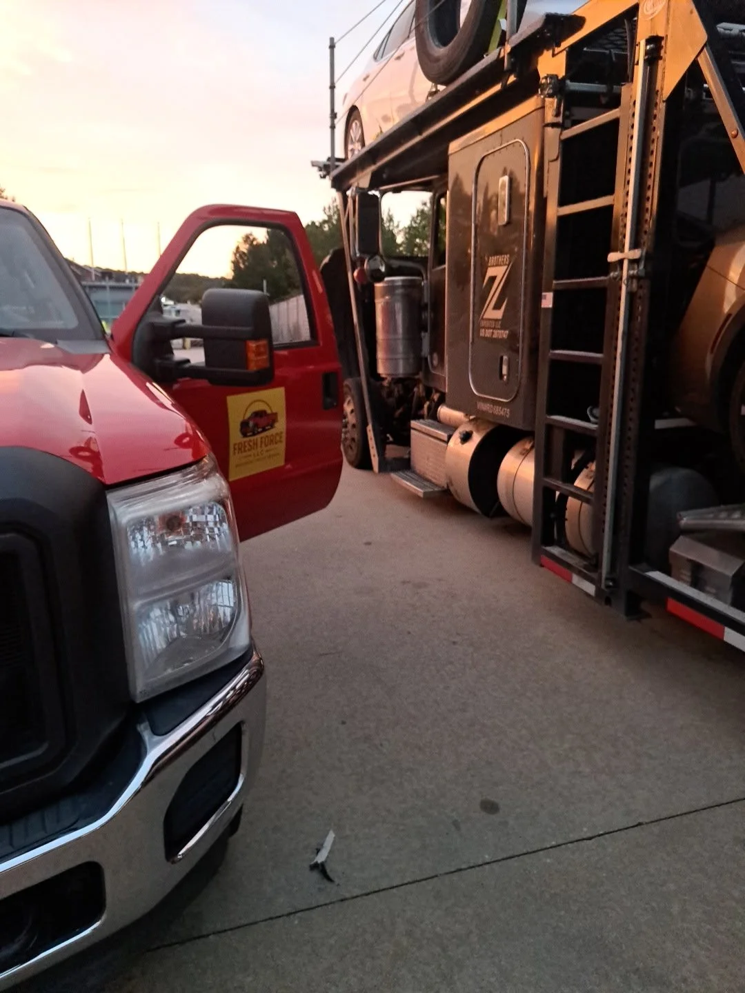 A red and a black vehicle, with the black vehicle's front partially visible, parked on a concrete surface with a food truck in the background and a sunset sky.