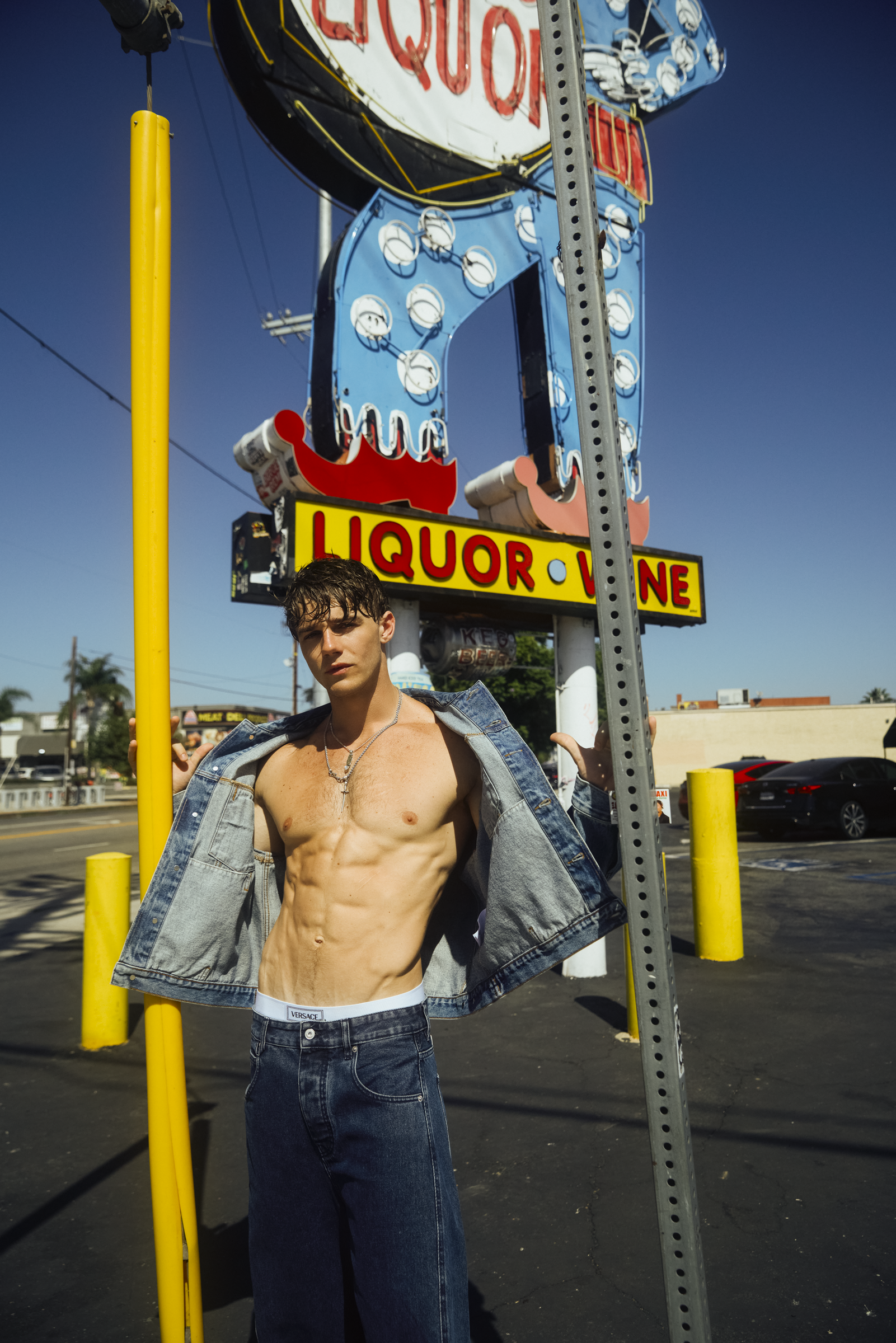 A young man with dark, wet hair standing shirtless outdoors, holding an open denim jacket, in front of a parking lot with yellow poles, holding onto a metal pole, with vintage neon signs for a liquor store and a neon sign displaying part of a fast fo