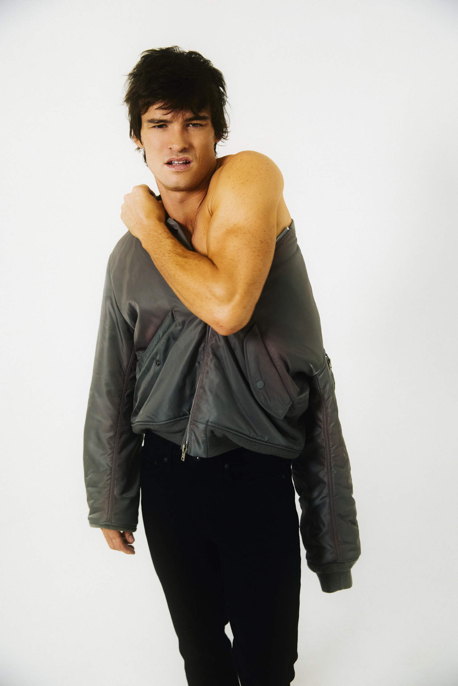 A young man with dark hair, wearing a gray jacket and black pants, standing against a plain white background. He appears to be stretching or shrugging, with one arm crossed over his chest and the other hanging loosely by his side.