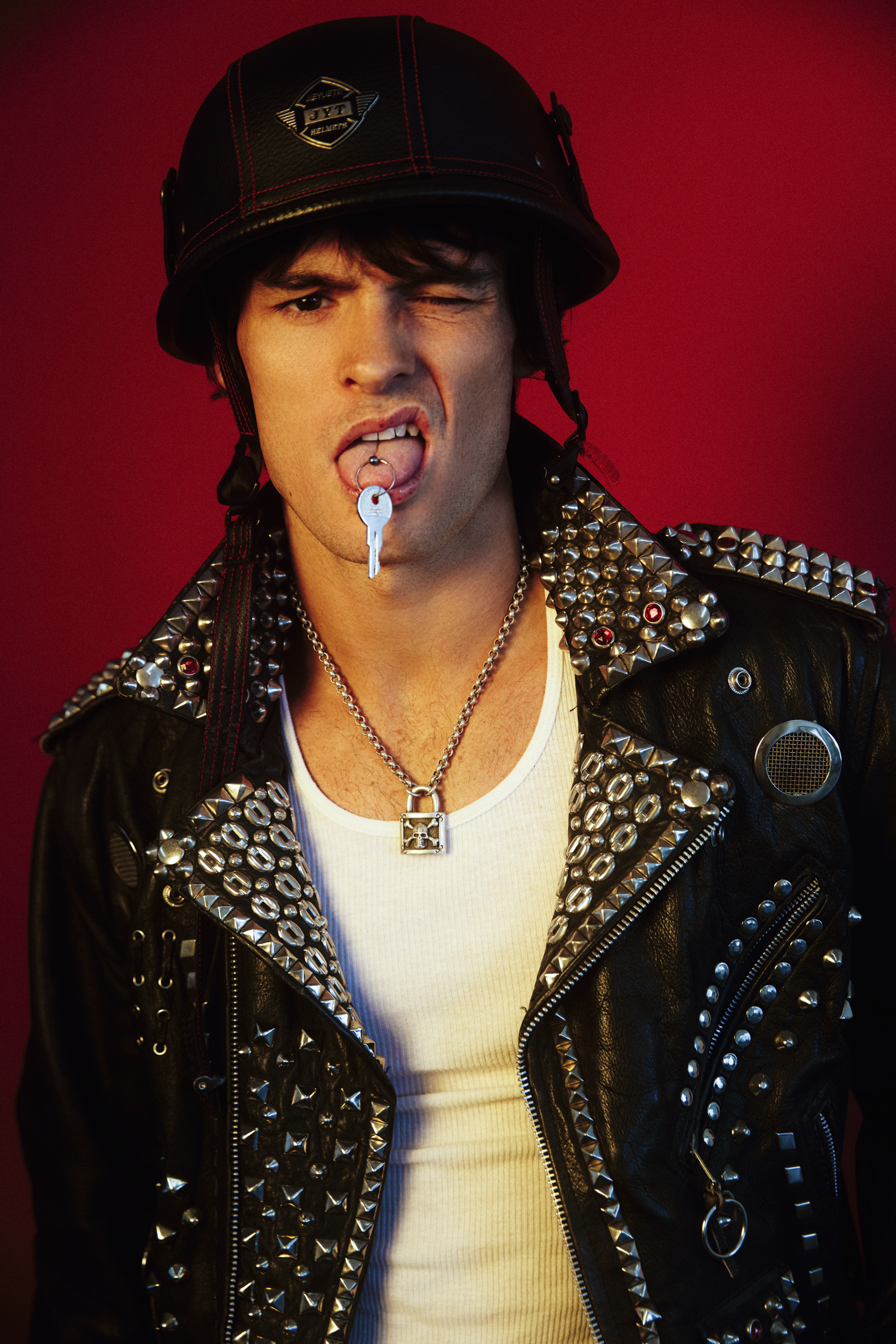 Young man wearing a black leather jacket with silver spikes and studs, a white shirt, a black helmet with red stitching, a key pendant necklace, and a dog tag hanging from his tongue, against a red background. He is winking with one eye and has a pie