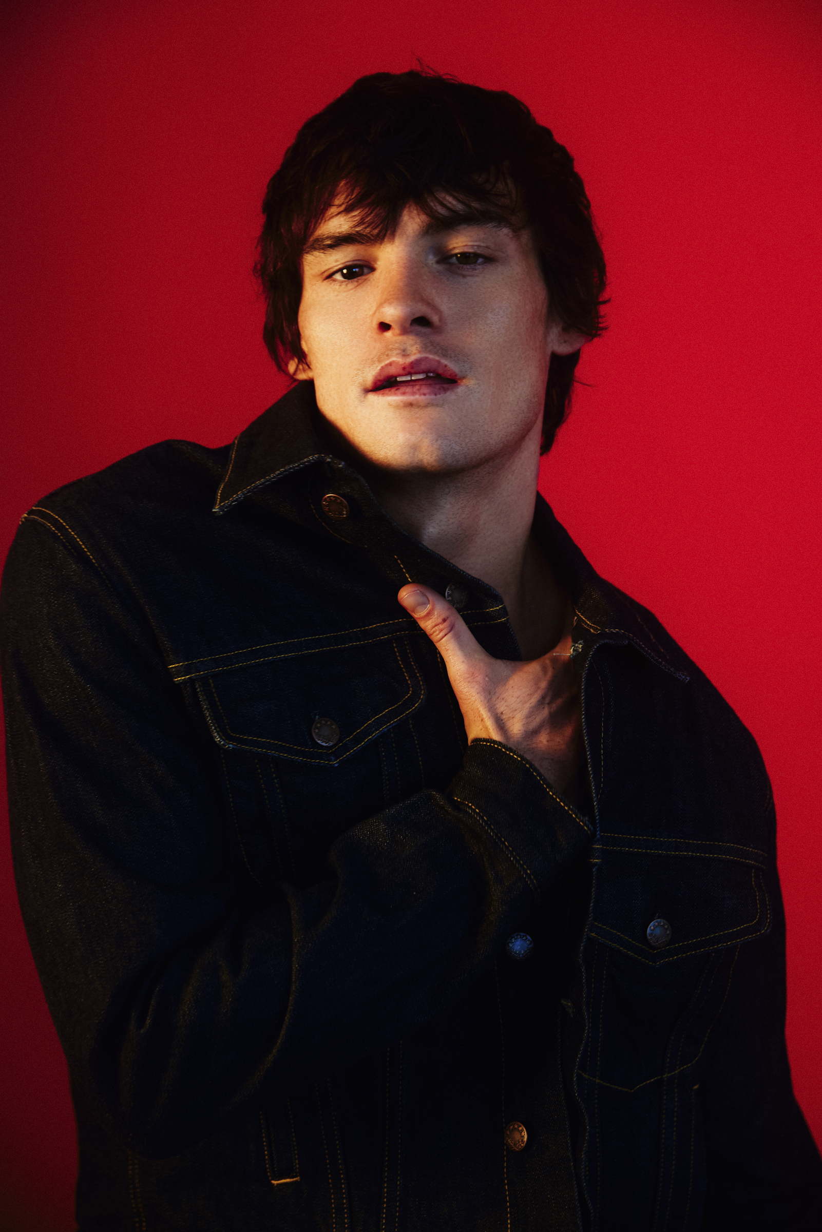Young man with dark hair and a denim jacket against a red background, slightly biting his lip.