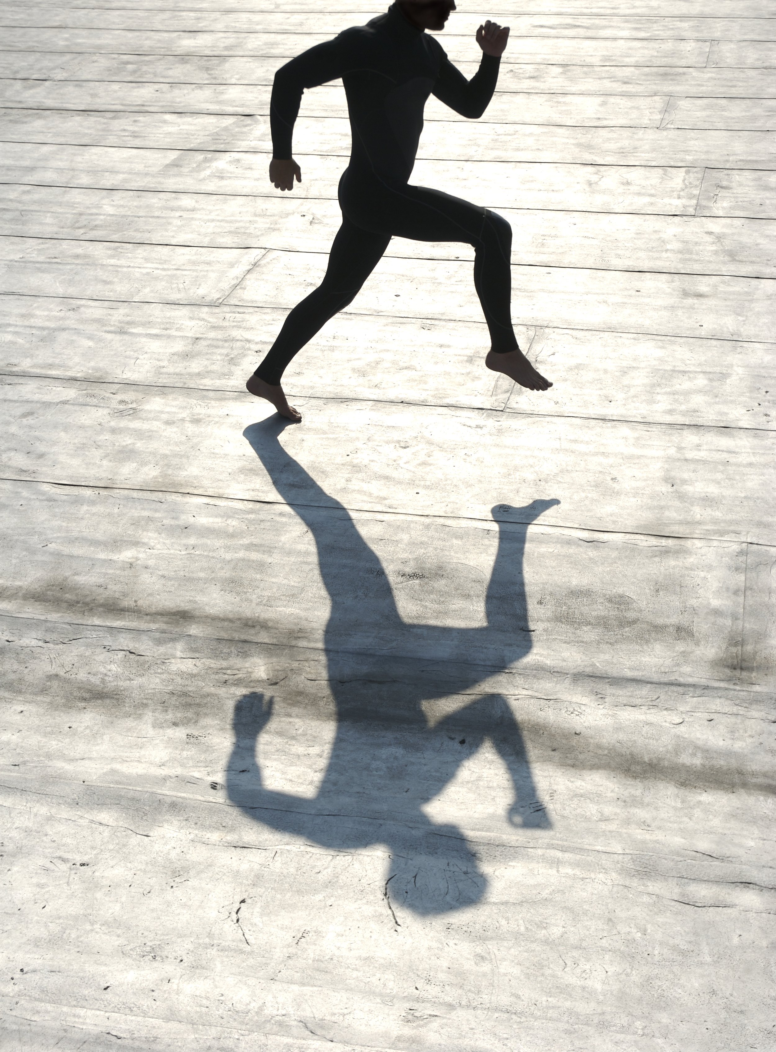 Silhouette of a person running or jogging on a light-colored wooden surface, with their shadow cast on the ground.