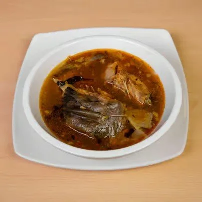 A bowl of fish stew with fish and vegetables in a broth, placed on a white dish on a wooden surface.